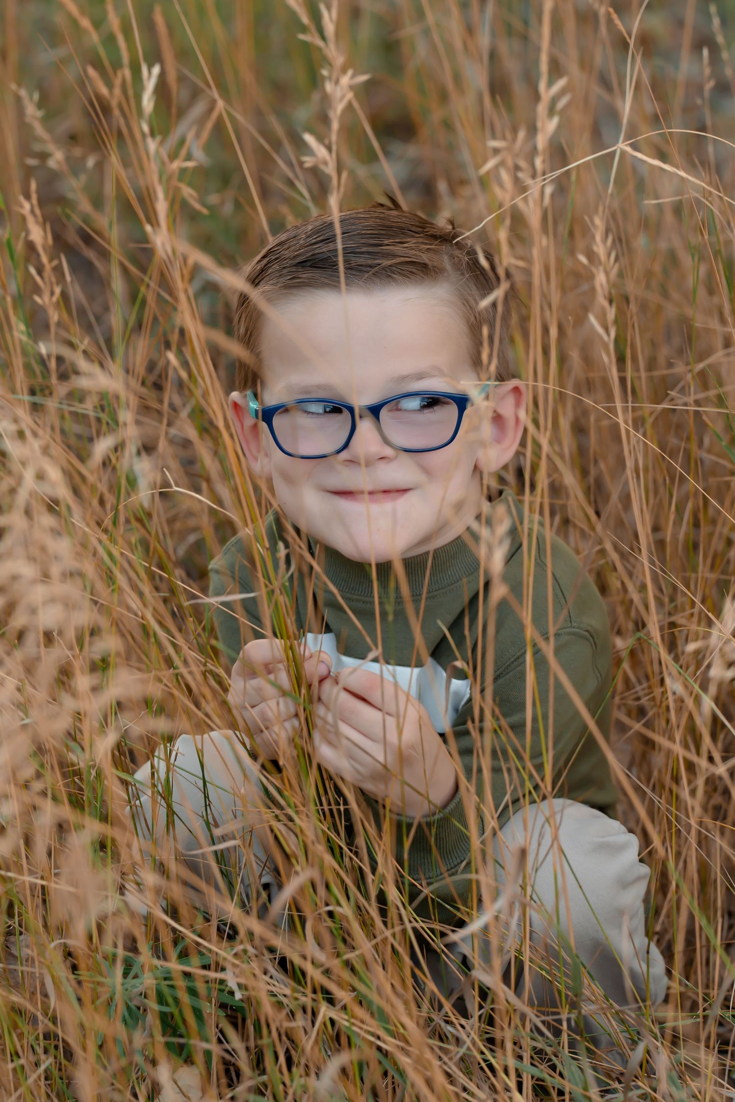 Young person with glasses peeks through tall autumn grass during outdoor portrait session.