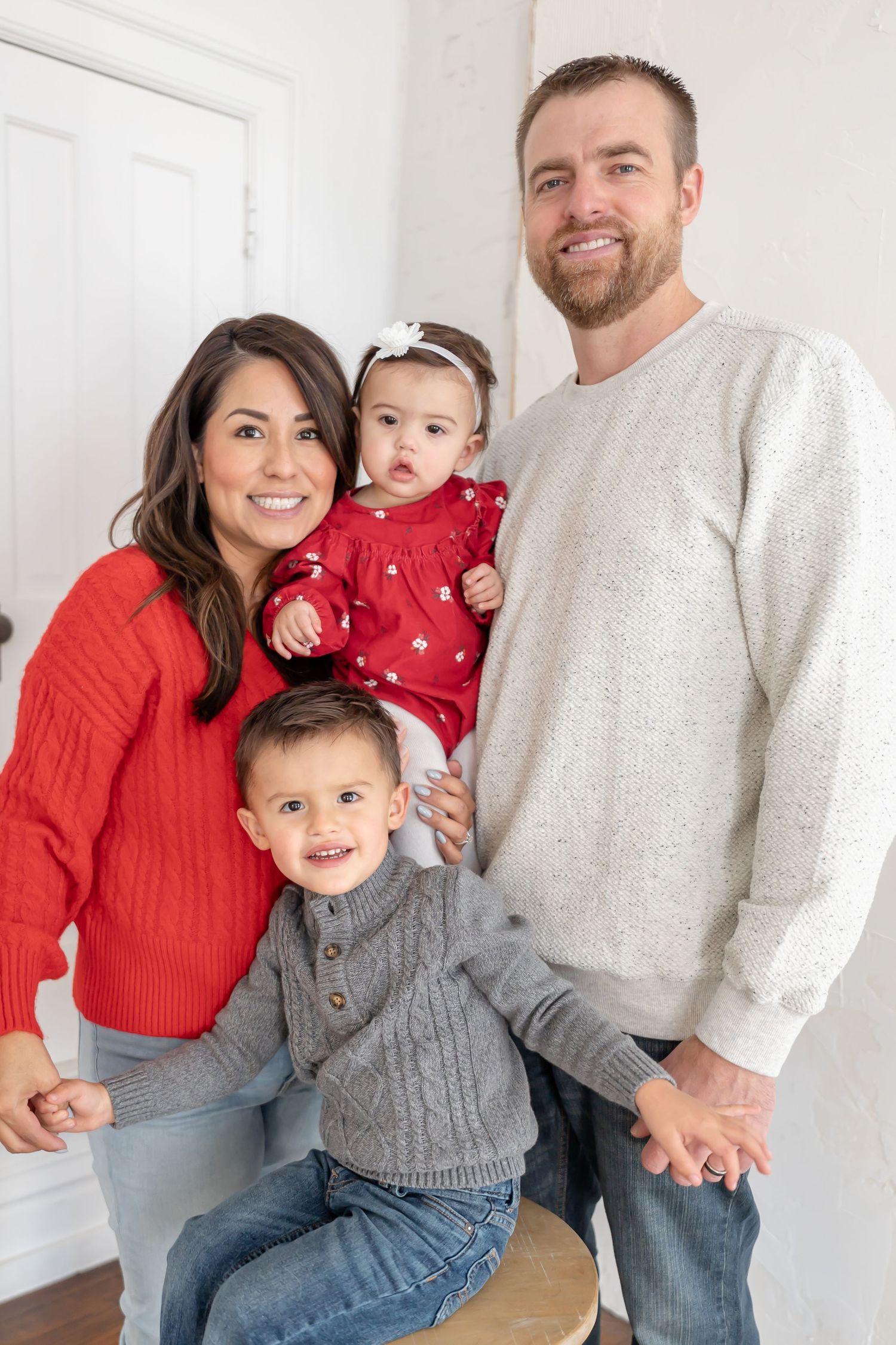 Family wearing coordinating red and gray sweaters poses together for holiday photos against white wall.