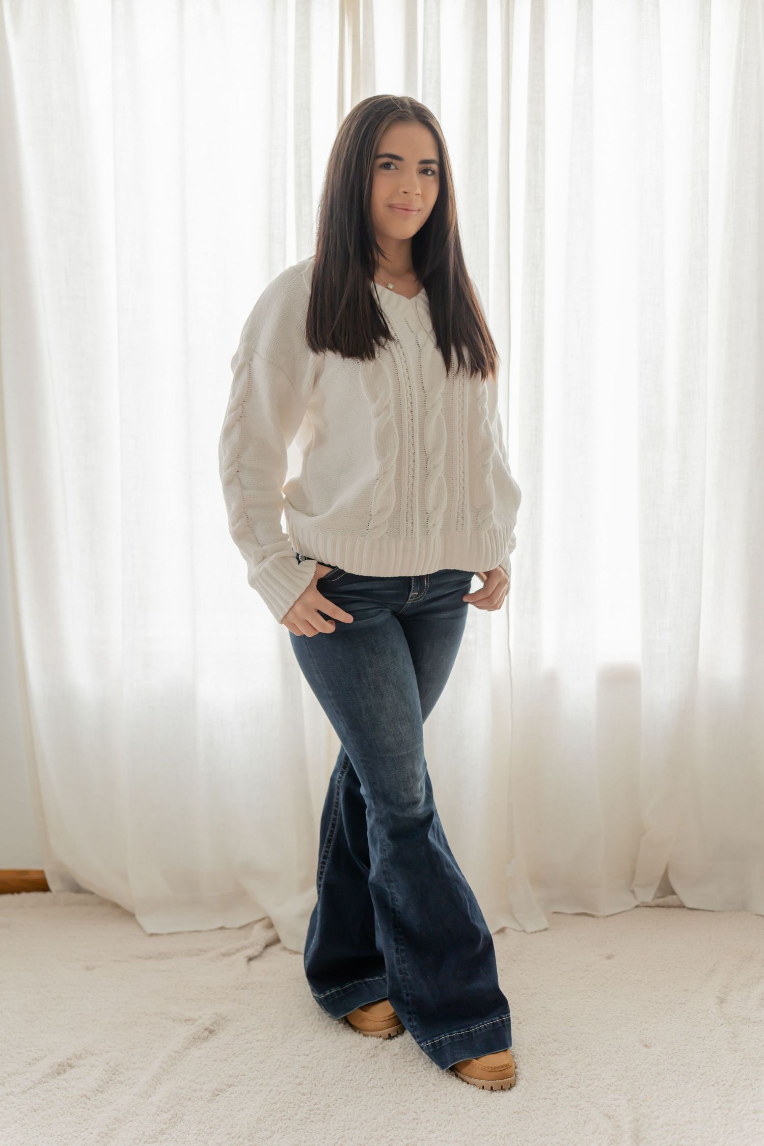 Model in white sweater and flared jeans poses against white curtain background in fashion photoshoot series.