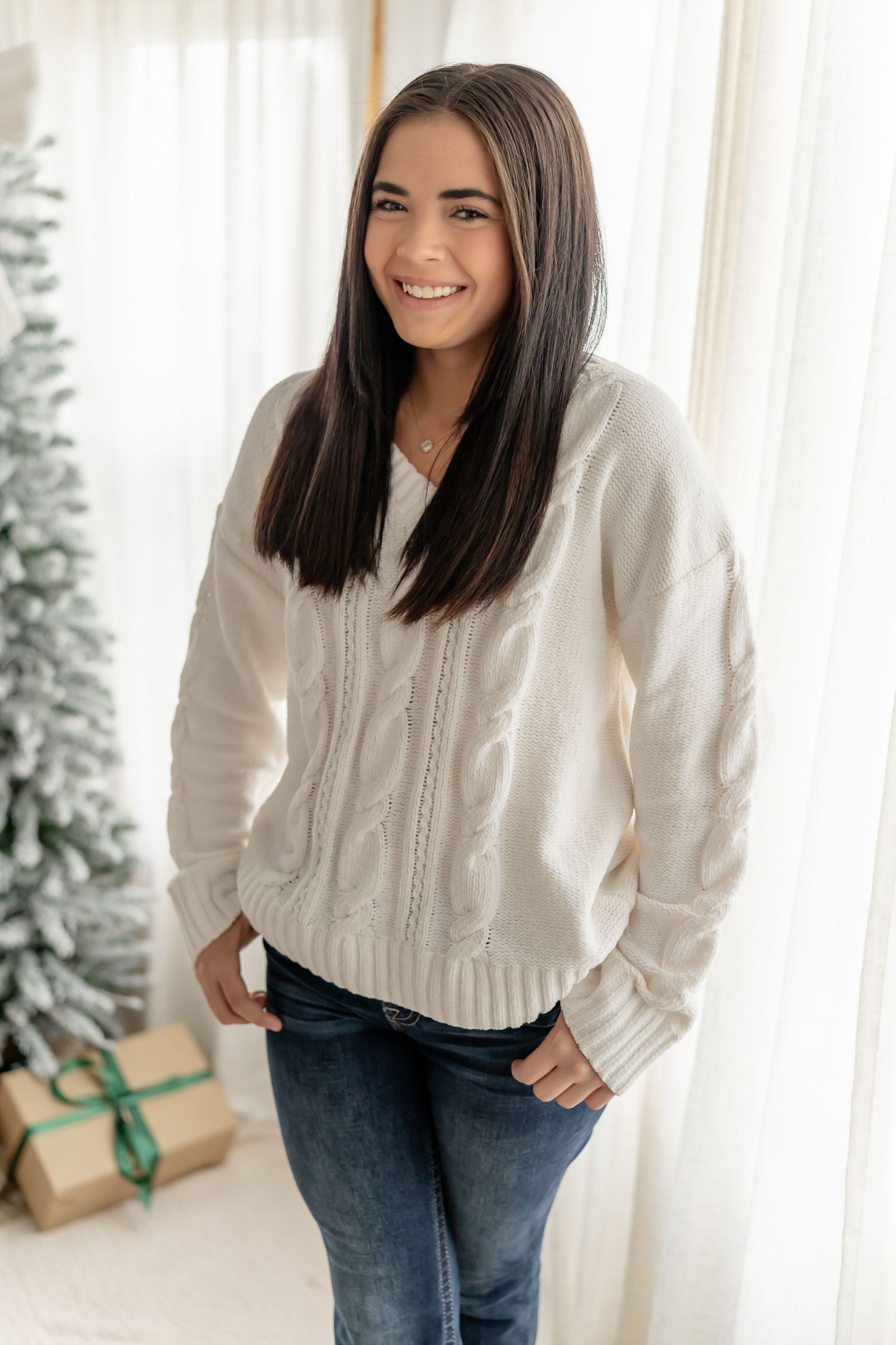 A person wearing a white sweater and jeans poses in front of a bright window with holiday decorations visible.
