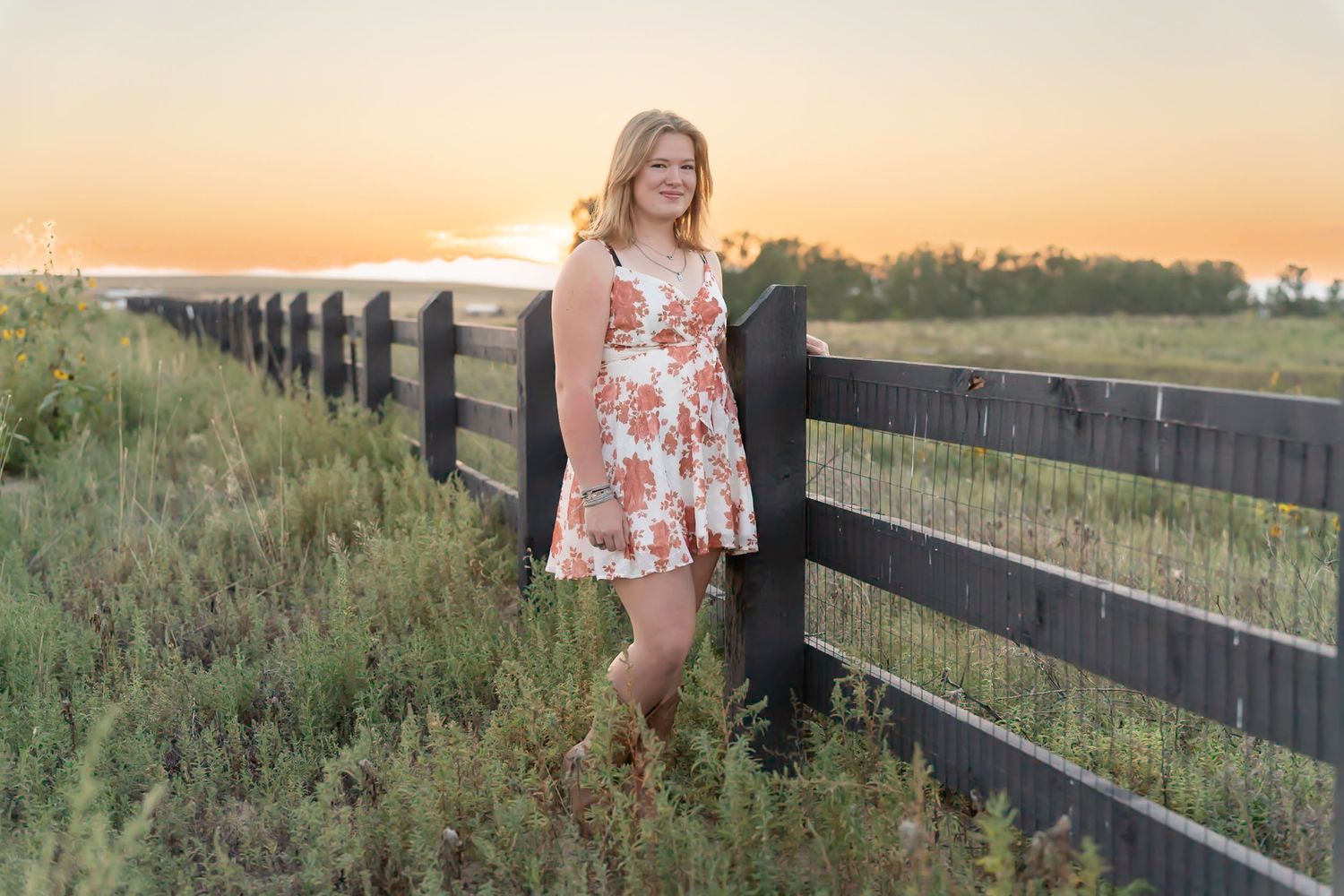 Woman in floral dress poses along wooden fence at sunset in rural field with golden light.