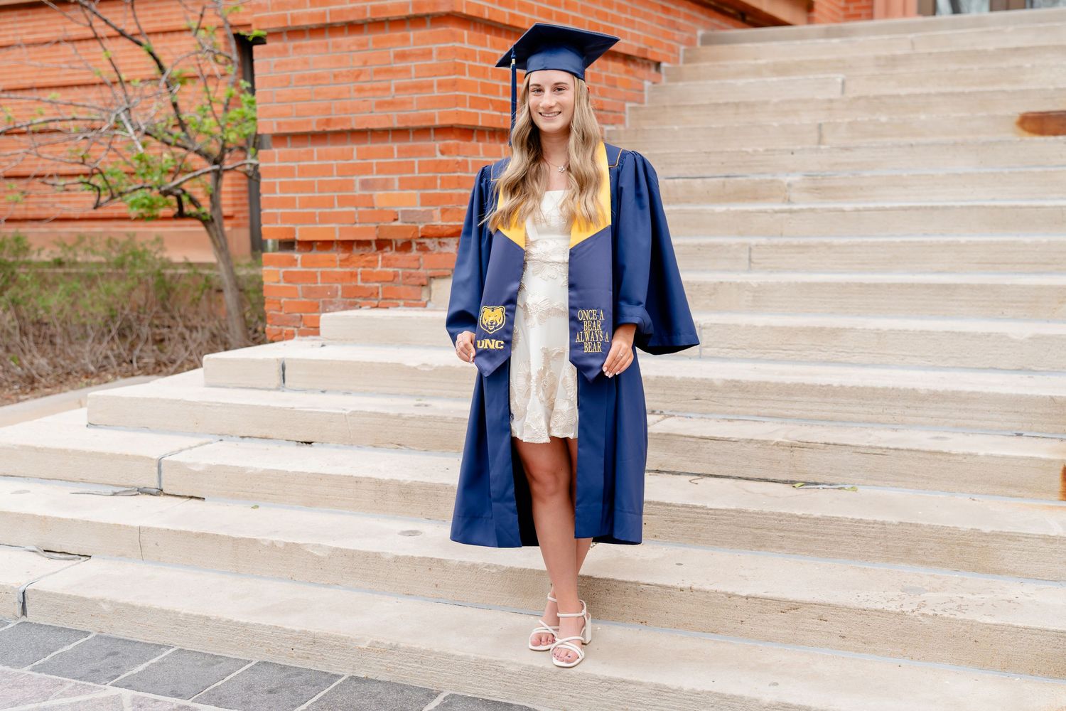 Graduate in blue cap and gown poses on concrete steps of a brick building.