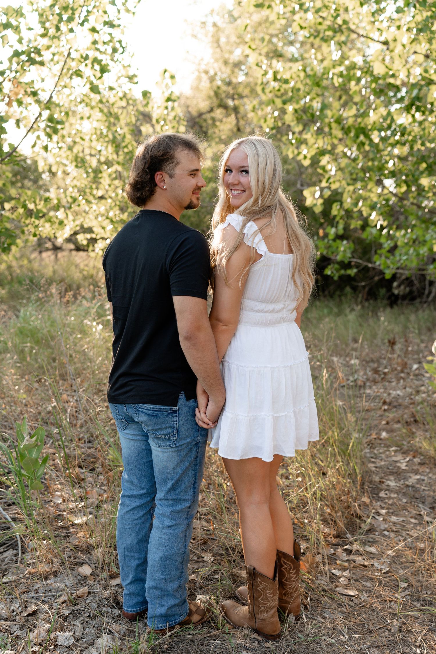 A couple in casual attire poses together outdoors among trees and tall grass.