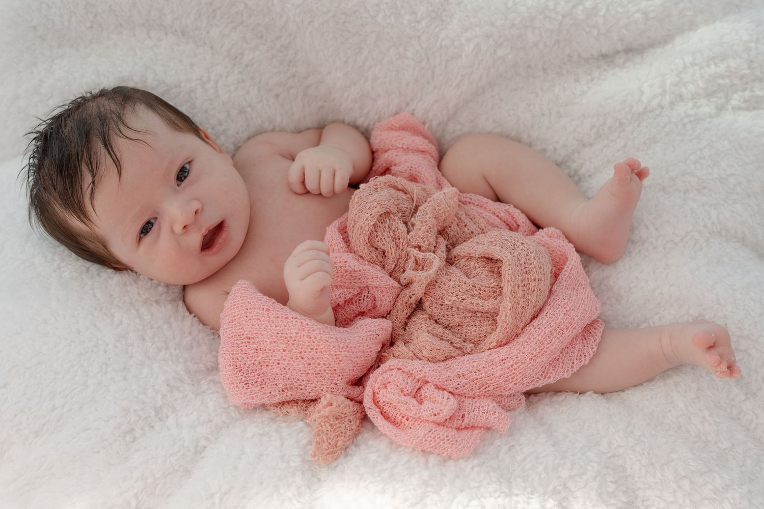 Baby lying on white blanket wearing coral pink outfit during professional photoshoot.