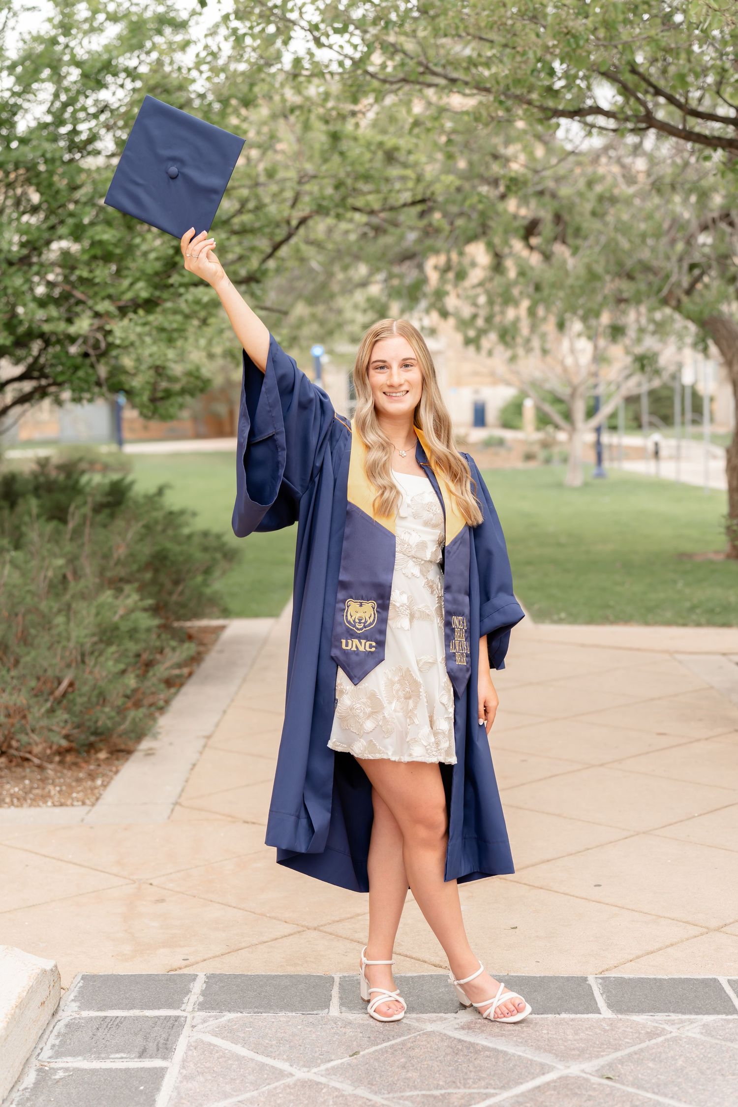 Graduate in blue cap and gown poses playfully outdoors on campus walkway.