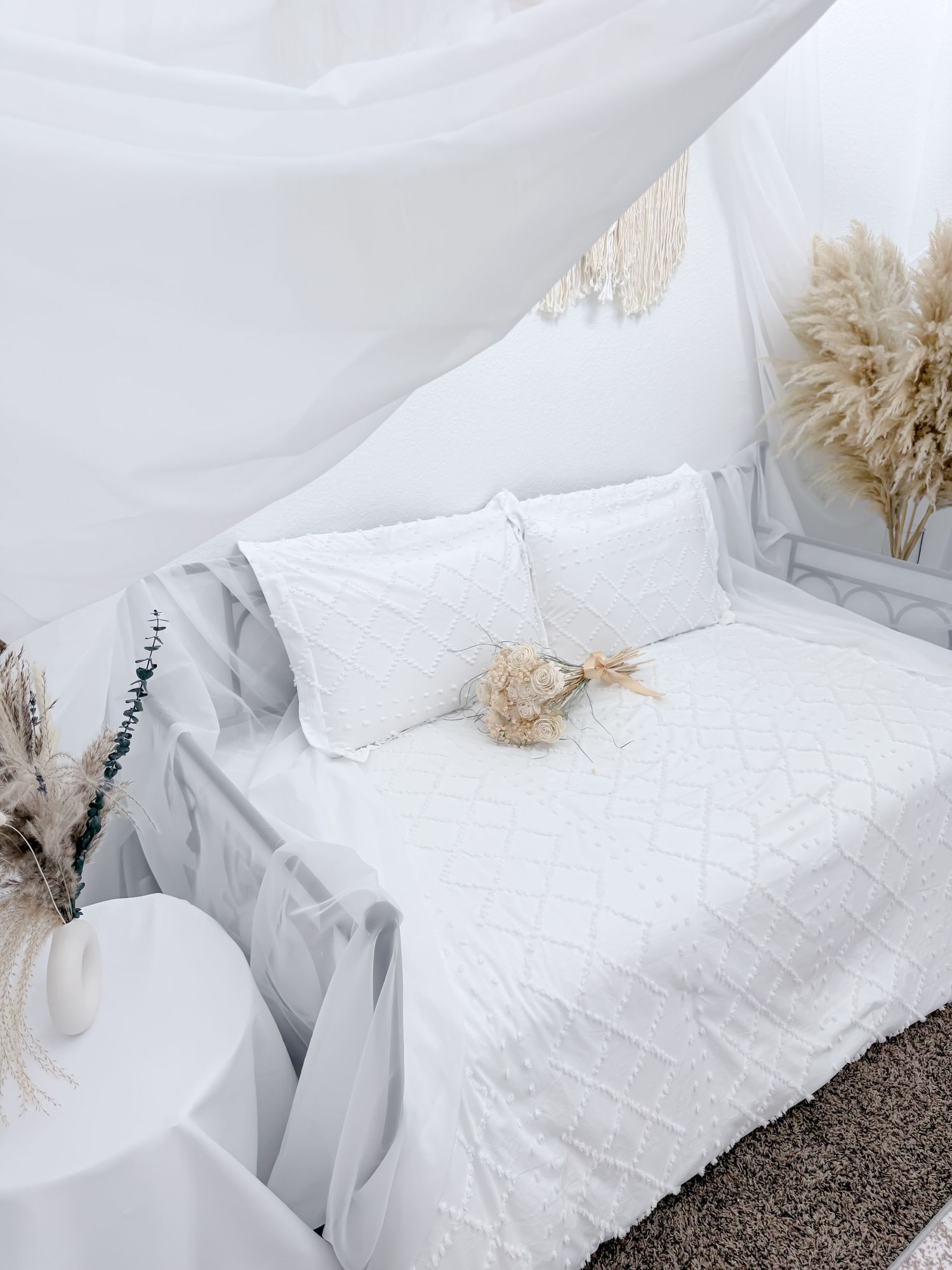 White linen bedding and textured throw pillows create a serene, minimalist bedroom setting with dried pampas grass accents.