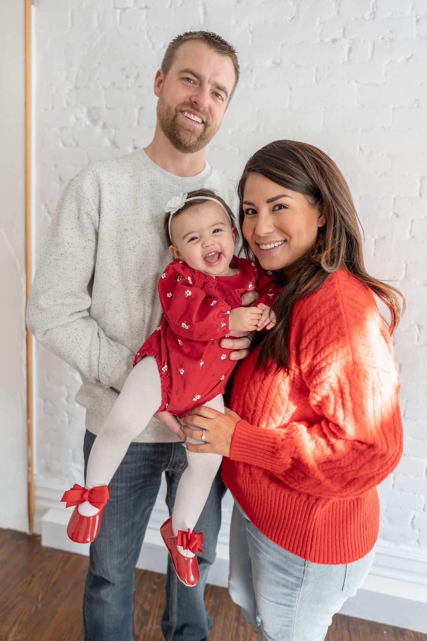 A family in coordinating red and gray outfits poses together against a white wall for holiday photos.