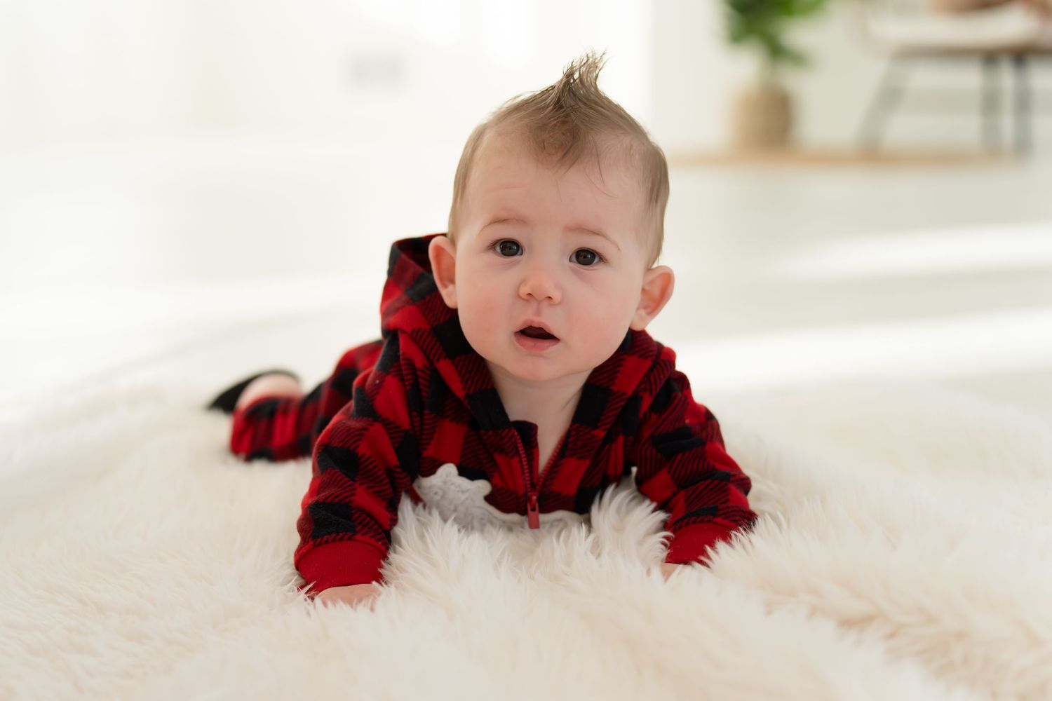 Baby wearing red and black buffalo plaid outfit crawls on white carpet while looking up at camera.
