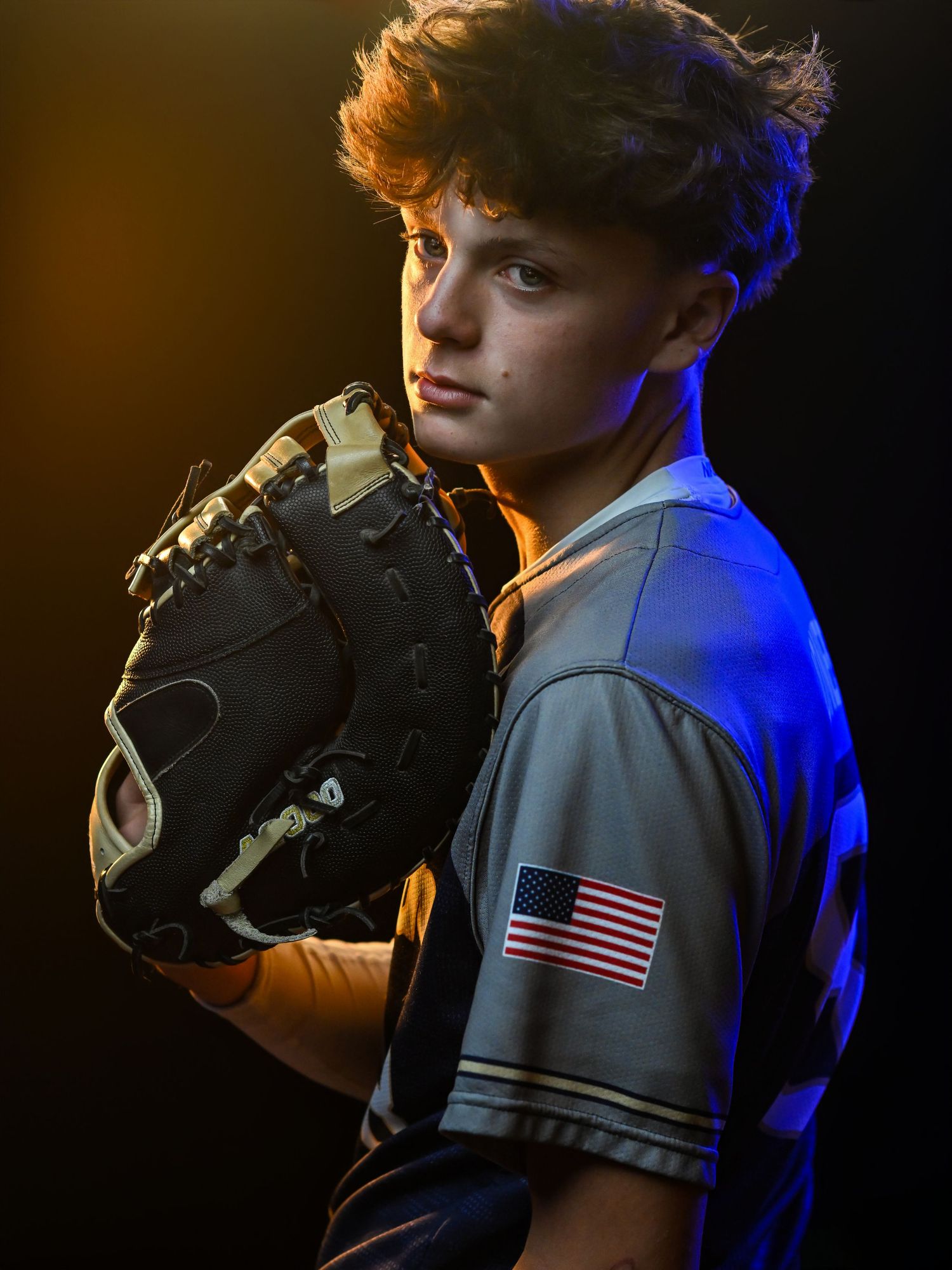 Baseball player in uniform with American flag patch poses with glove in dramatic lighting.