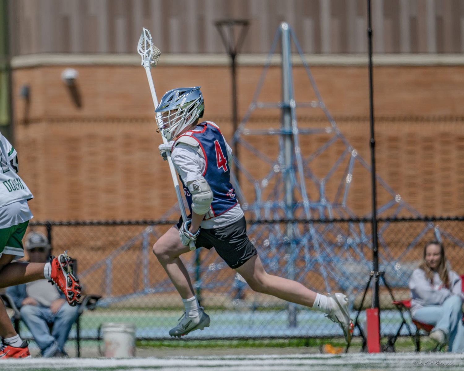 Lacrosse players' legs and feet lined up on artificial turf with stadium wall in background.