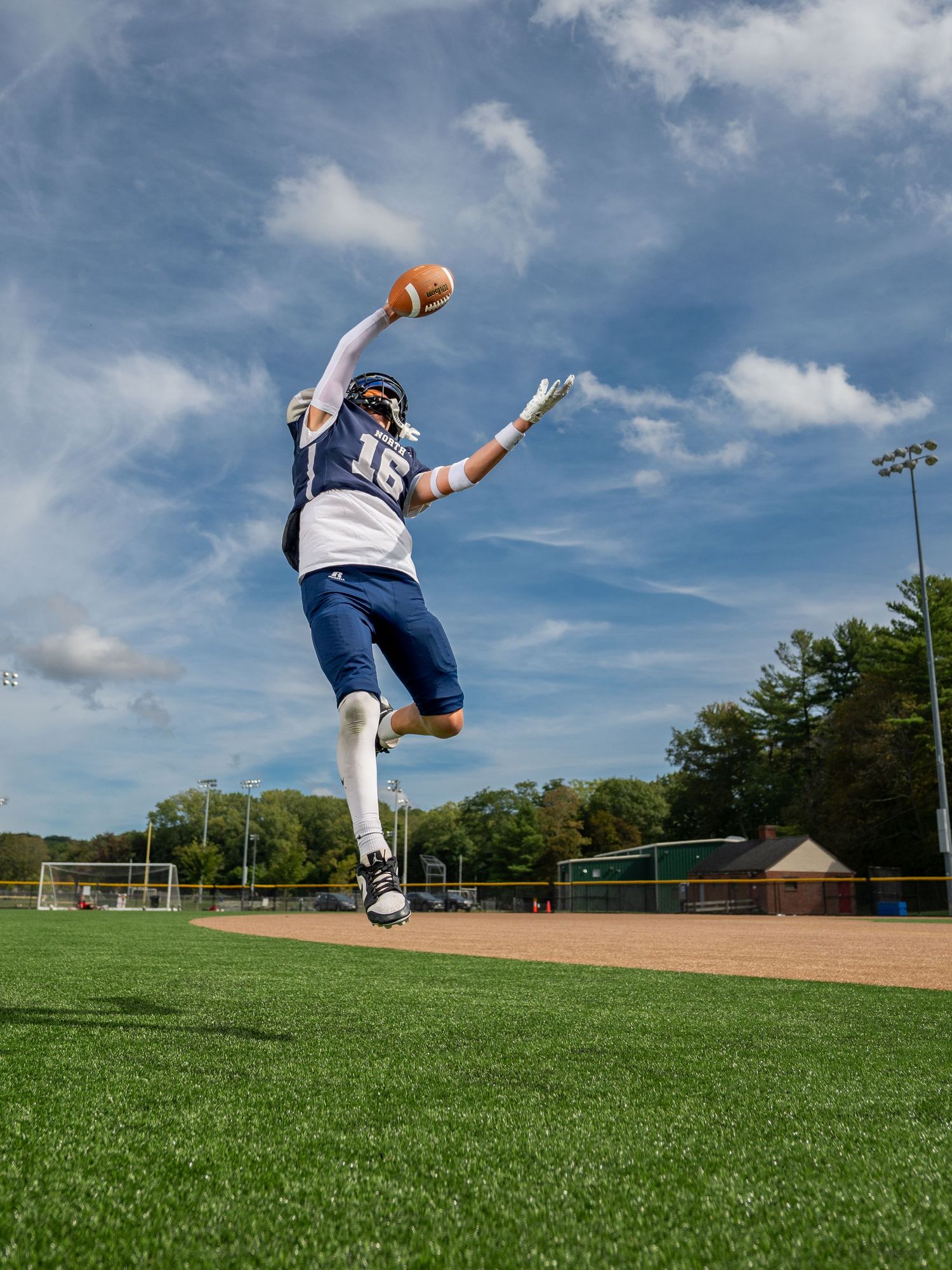 Football player leaping high in the air to catch a ball on a sunny day at an athletic field.