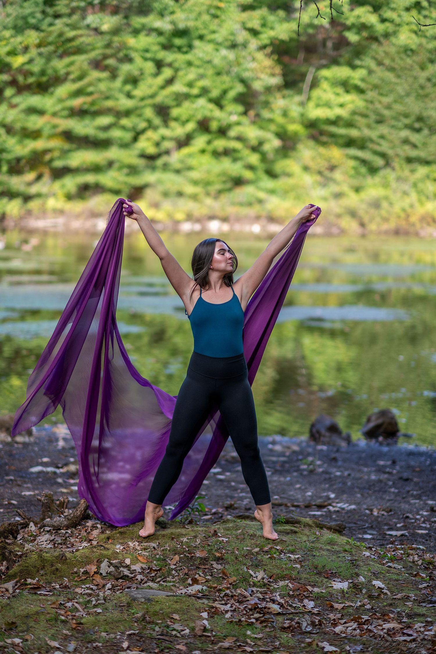 Dancer with purple fabric performs expressive movement by a lake surrounded by green foliage.