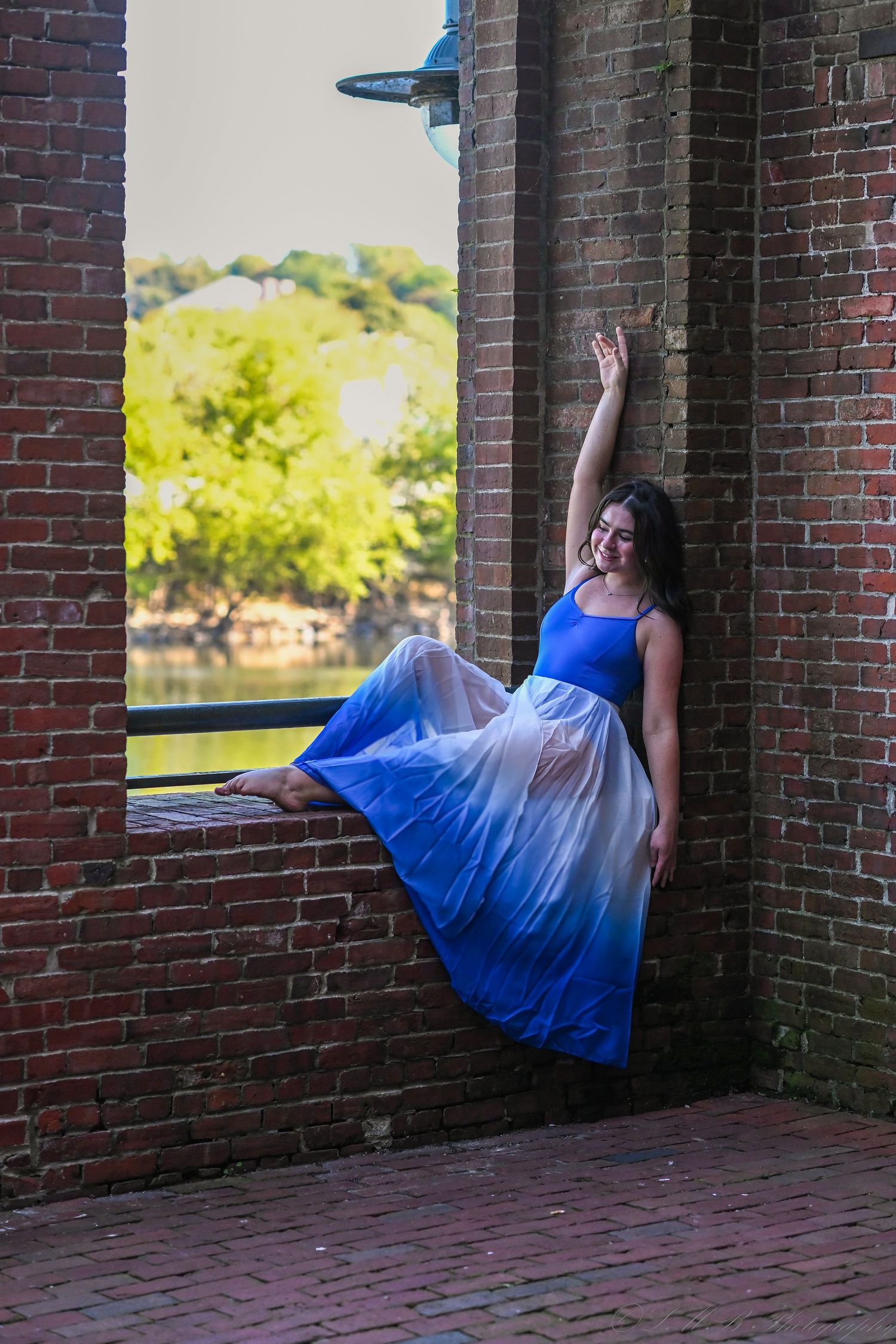 Dancer in blue ombre dress posing against brick archway with scenic background.
