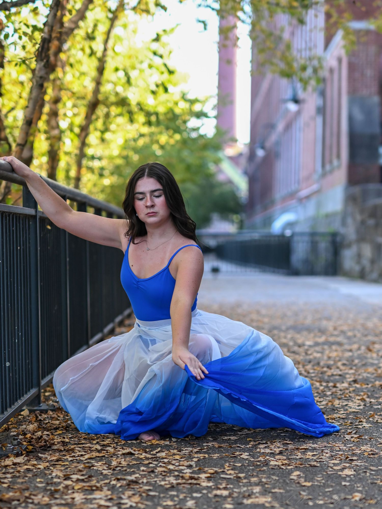 Dancer in blue and white tulle skirt poses gracefully on leaf-covered path.