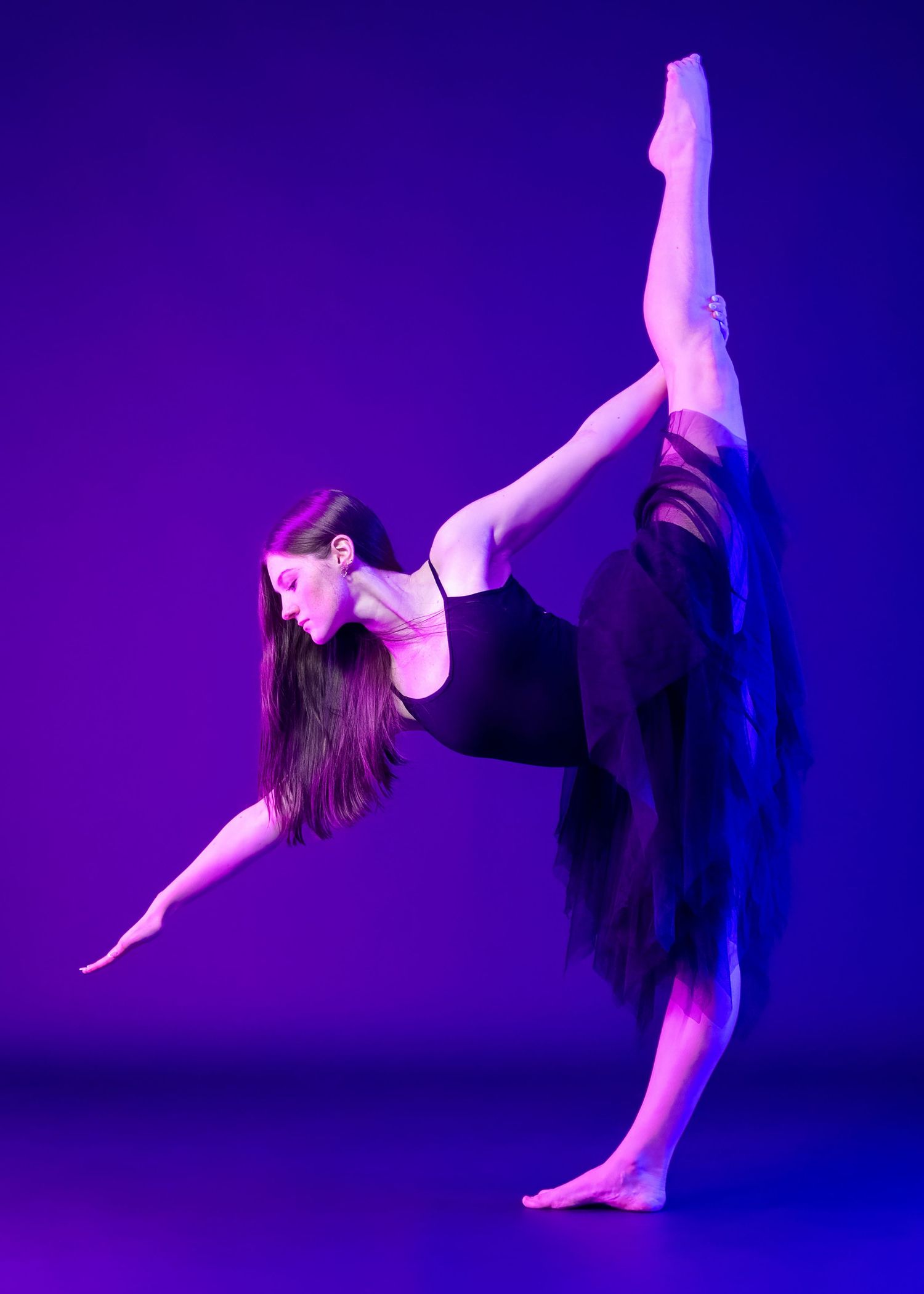 Dancer executes dramatic leg extension in purple-lit studio setting.