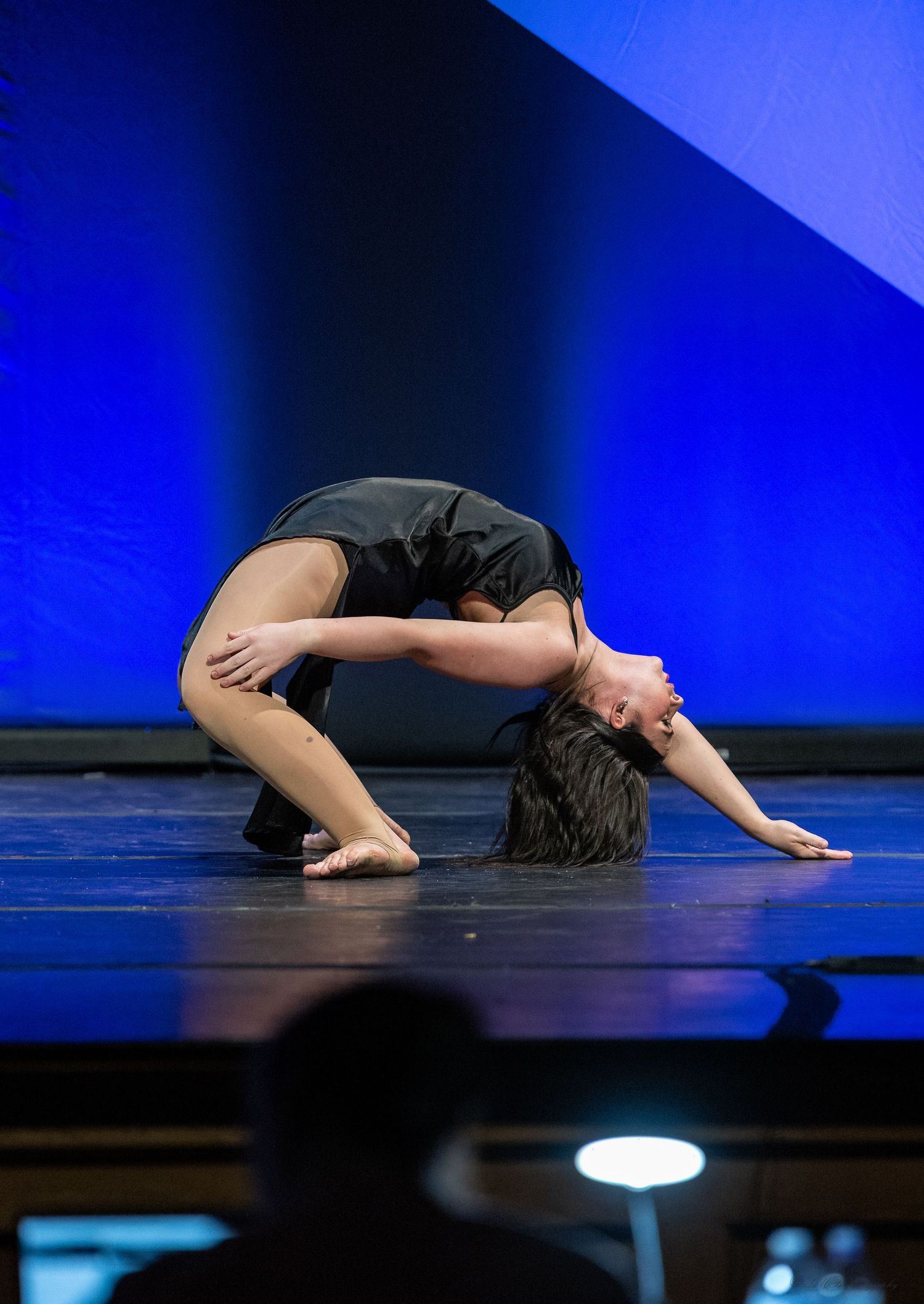 Contemporary dancer in black attire performs emotional floor routine on blue-lit stage.