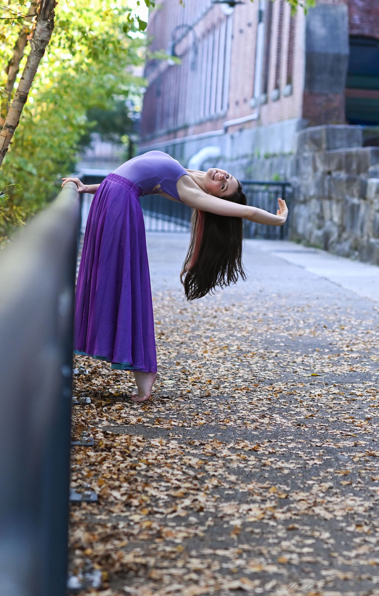 Dancer in purple dress arching backwards over railing on autumn path.