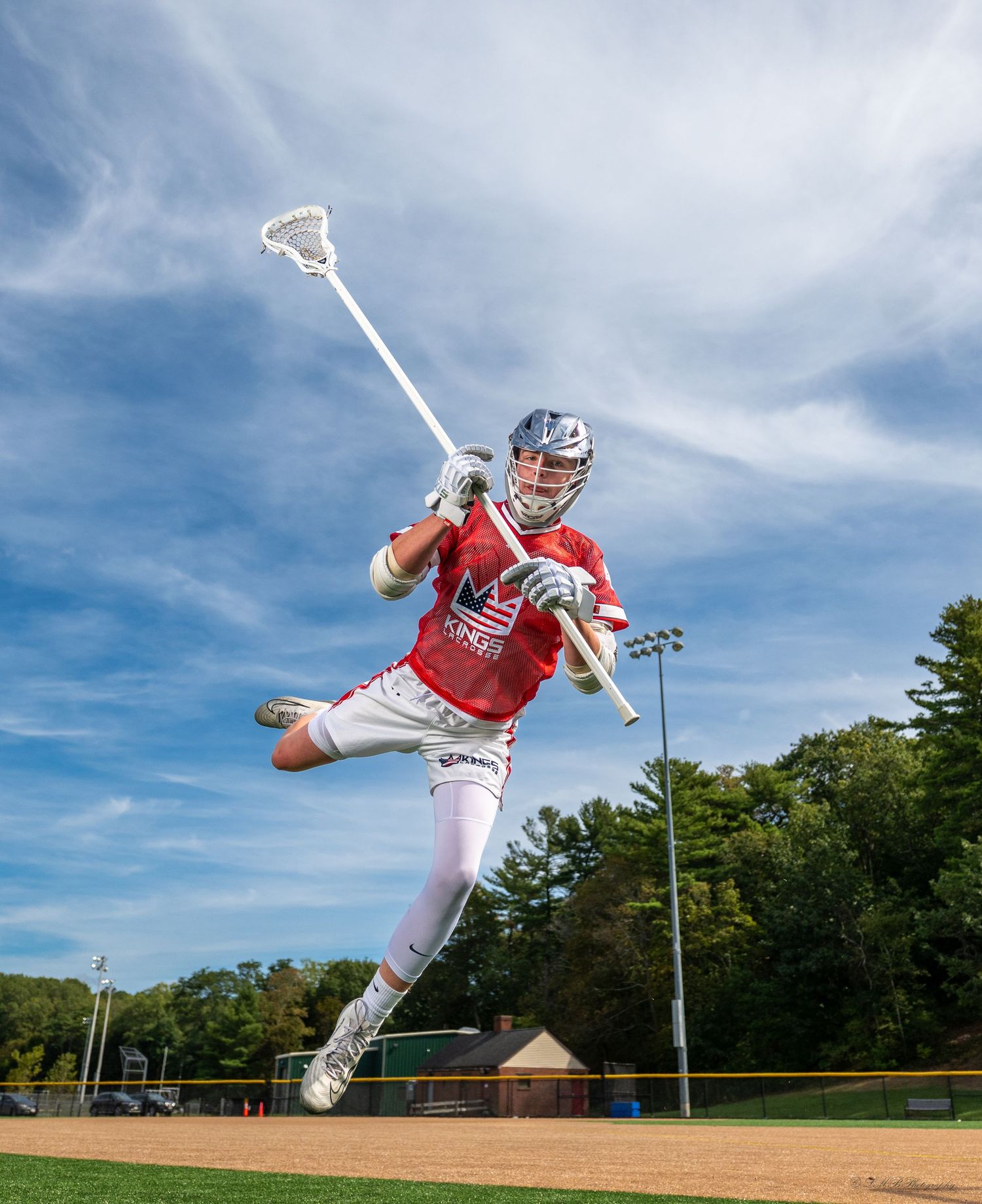 Lacrosse player jumping with stick extended against blue sky on sports field.