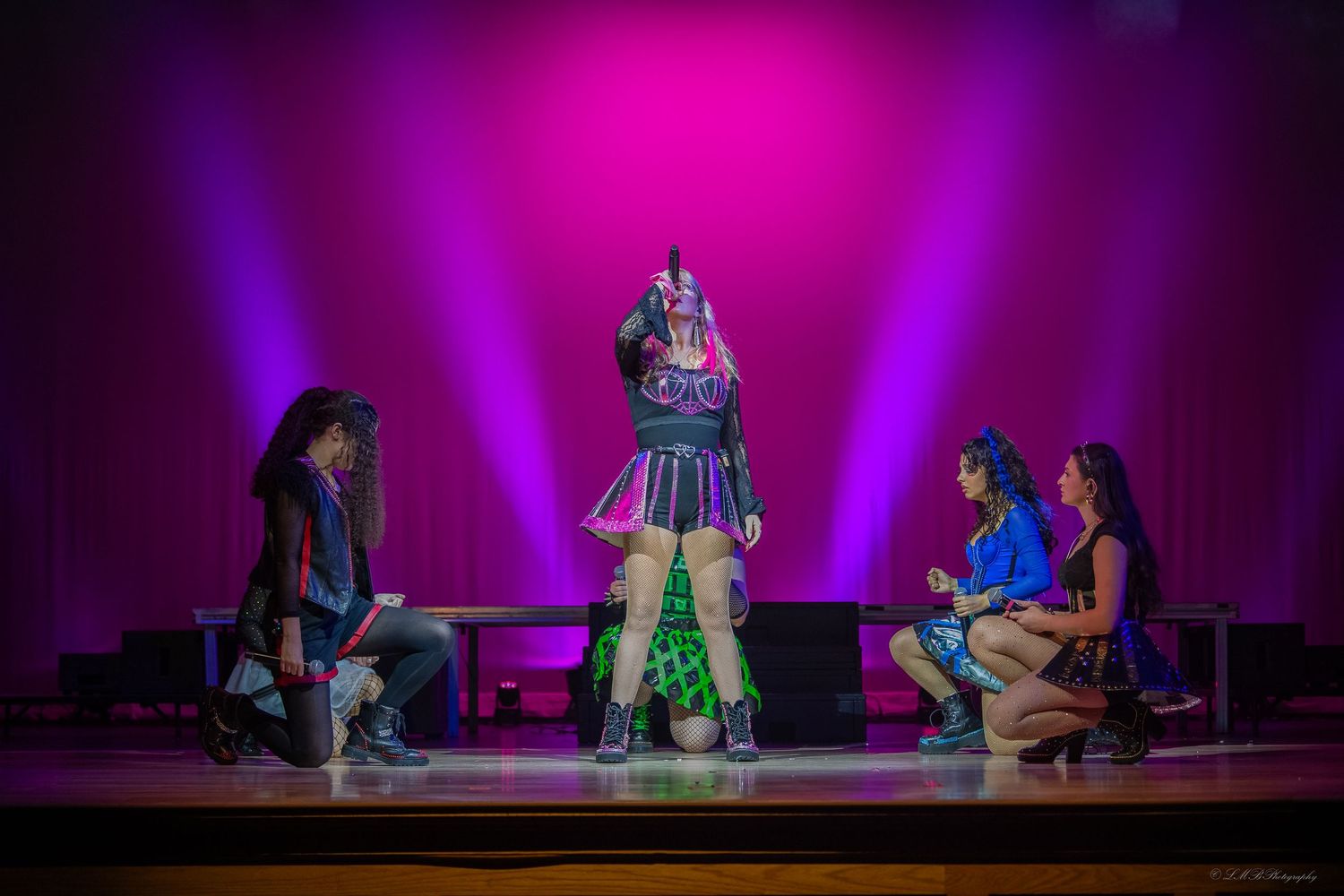 Theater performance on stage with vibrant magenta lighting and multiple performers.