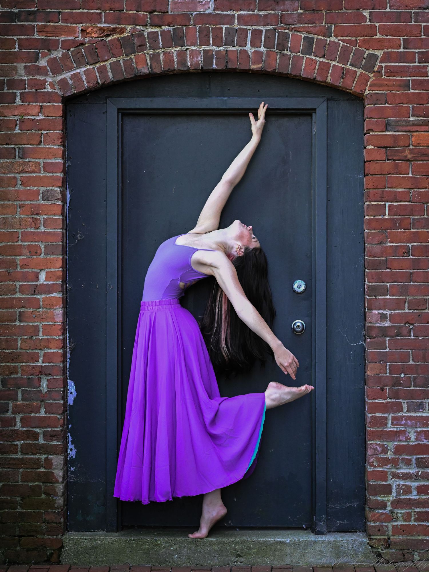 Dancer in purple dress performing elegant pose against dark door in brick archway.