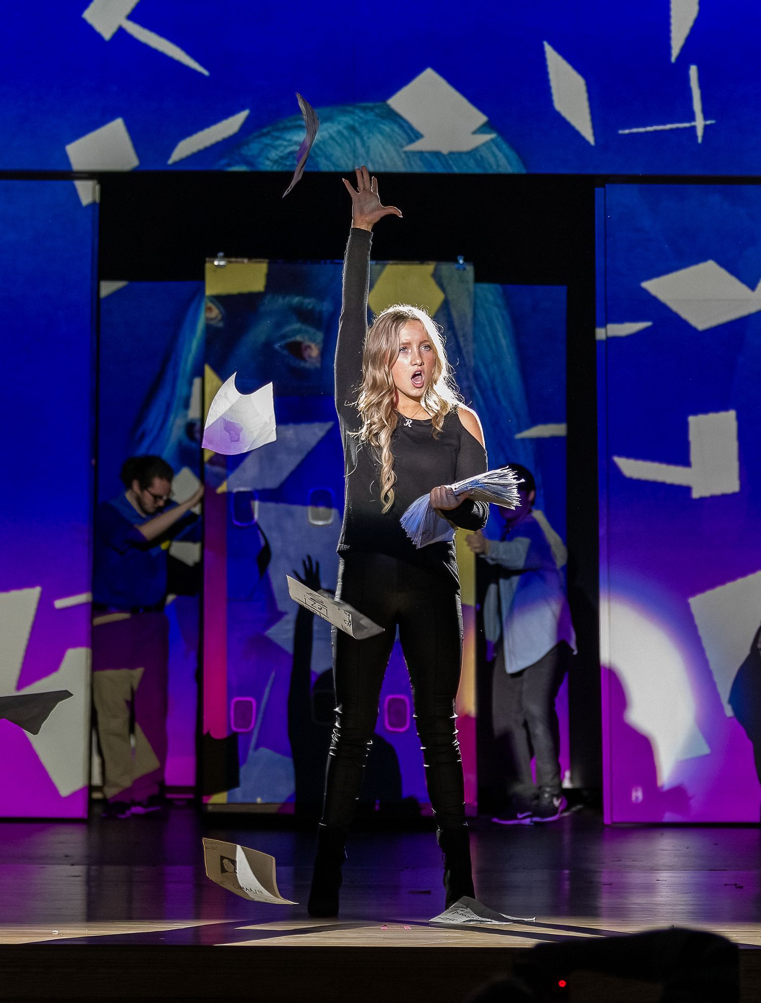 Stage performance with dramatic lighting featuring floating papers and artistic set design.