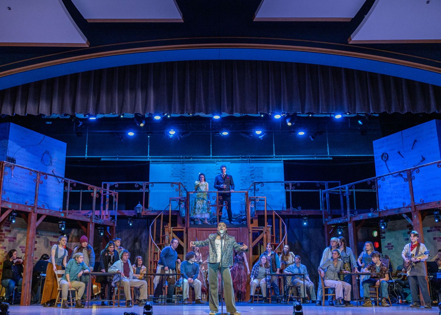 Wide theater stage with blue lighting and multiple performers arranged across the set.