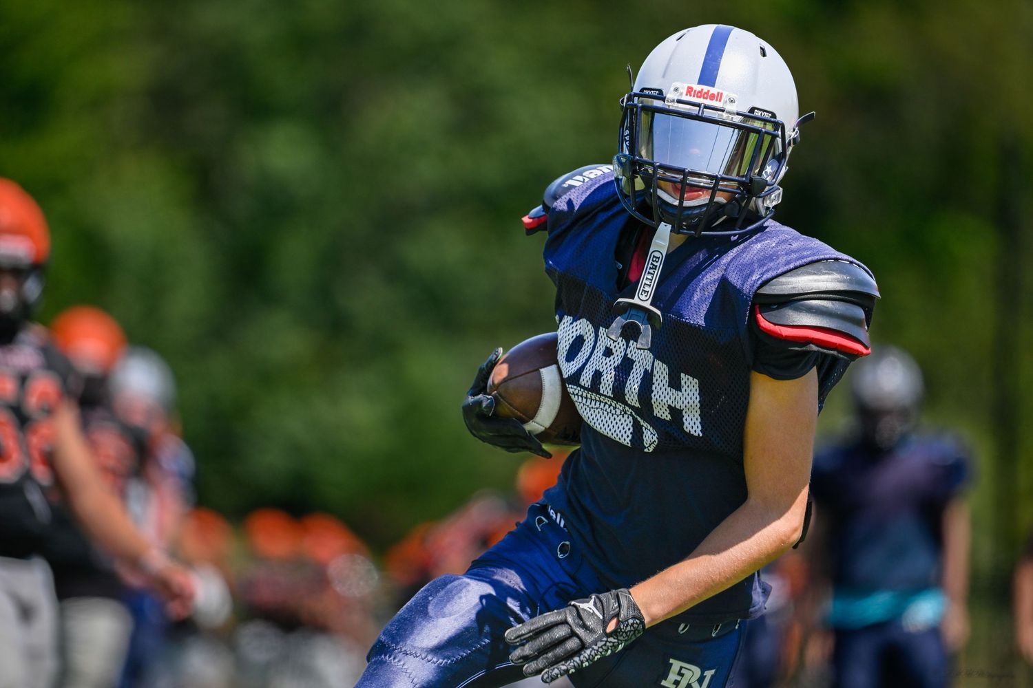 A football player in a dark blue uniform carries the ball during a sunny game.