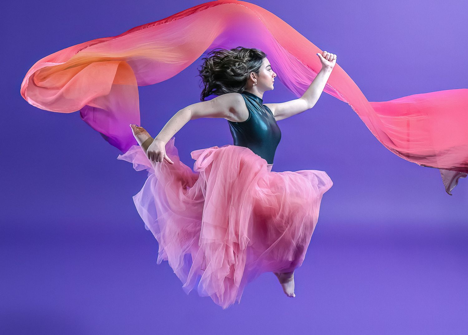 Dancer suspended mid-air wearing pink tulle skirt and black top with flowing coral fabric against purple background.