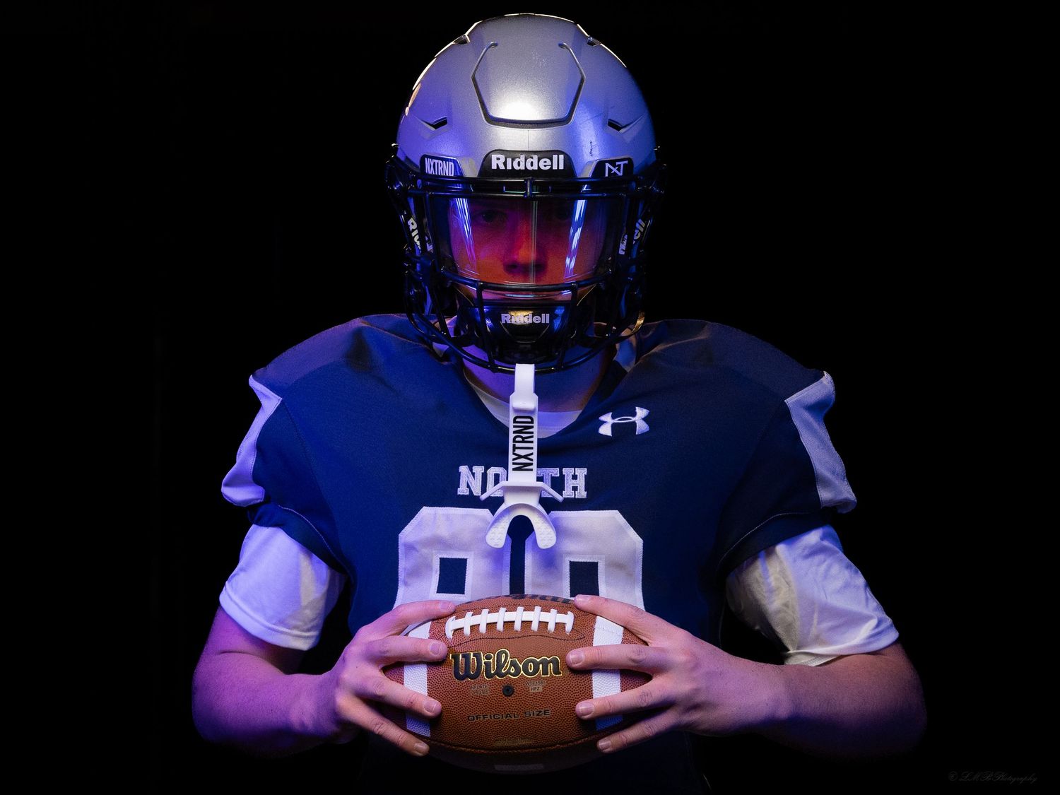 Dramatic studio portrait of football player holding ball in dark blue uniform with illuminated helmet.