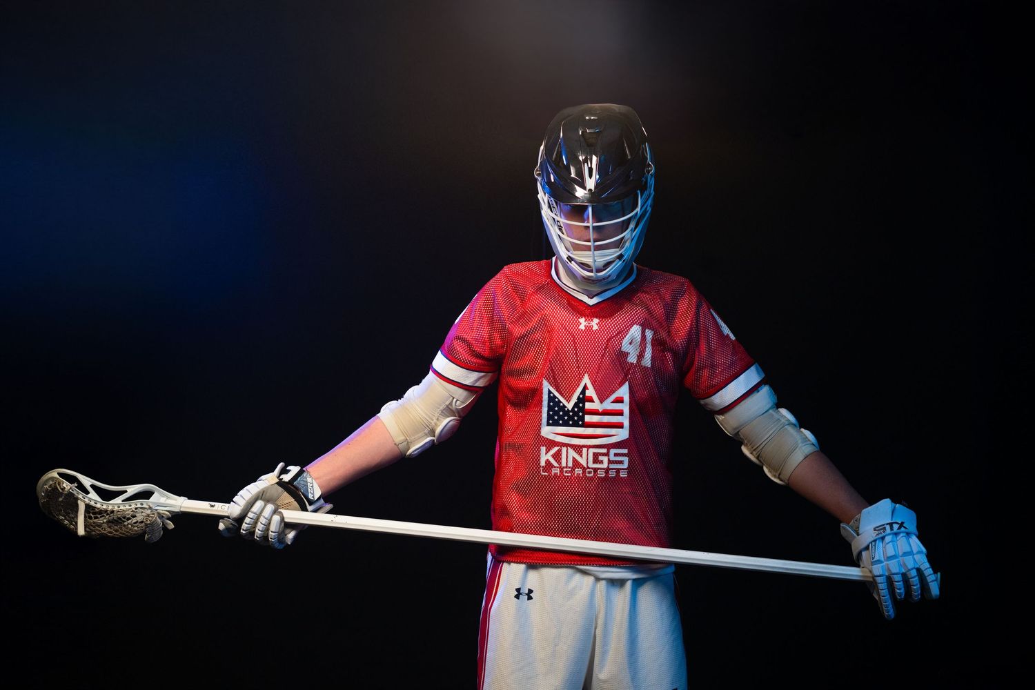 Lacrosse player in red jersey holding stick against dark background in dramatic lighting.