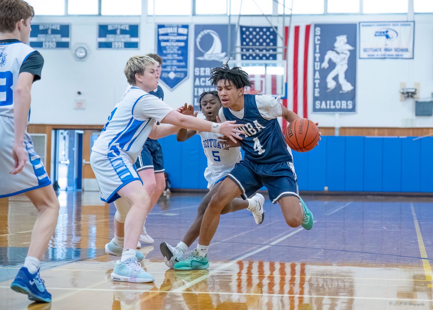 Basketball players compete for the ball on a glossy indoor court with American flag visible.