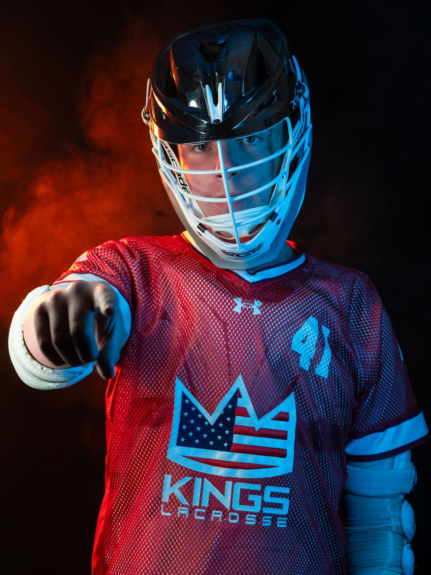 Lacrosse player in red jersey and helmet points forward against dramatic smoke background.