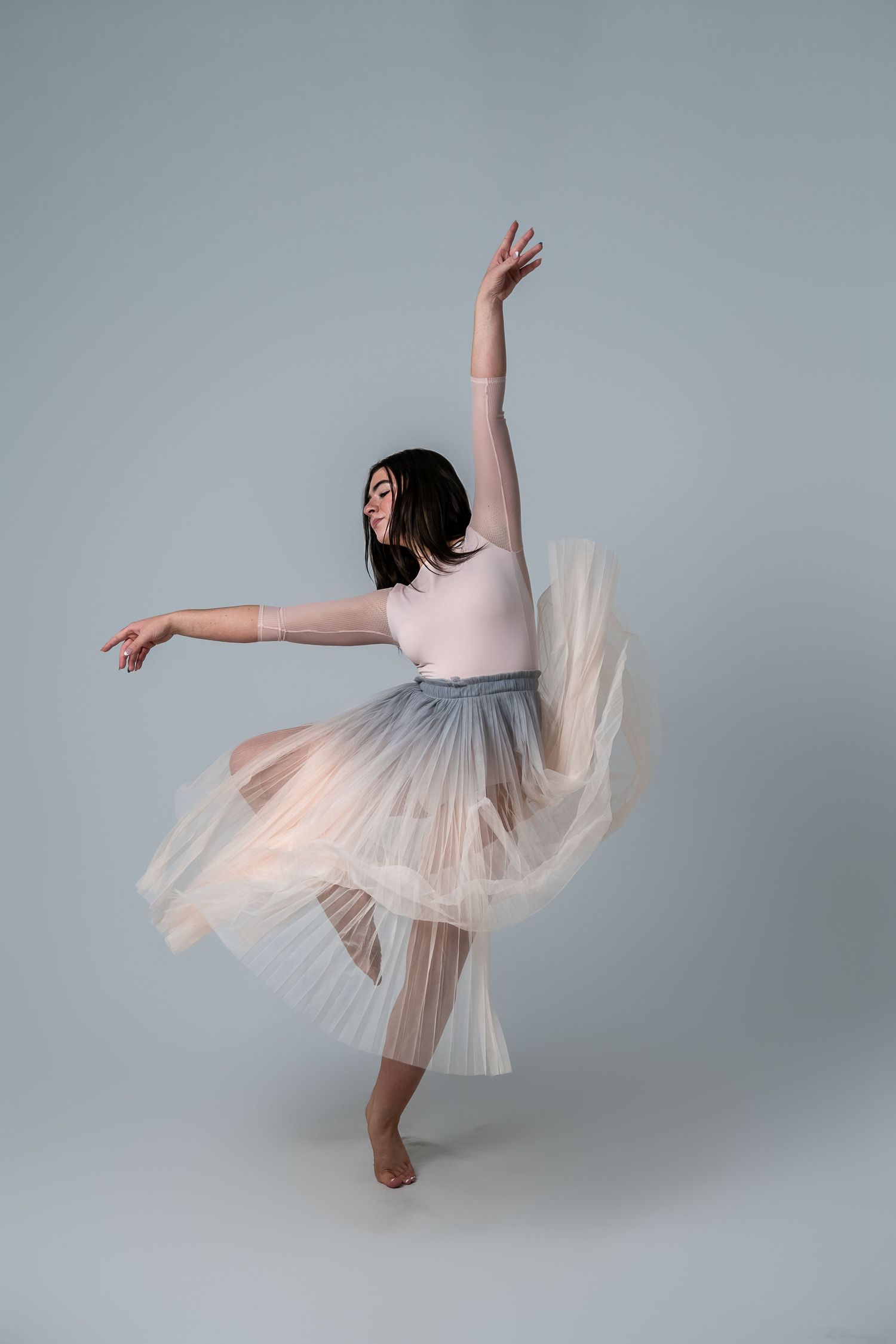 Dancer in pale pink tulle skirt performs graceful pose against grey background.