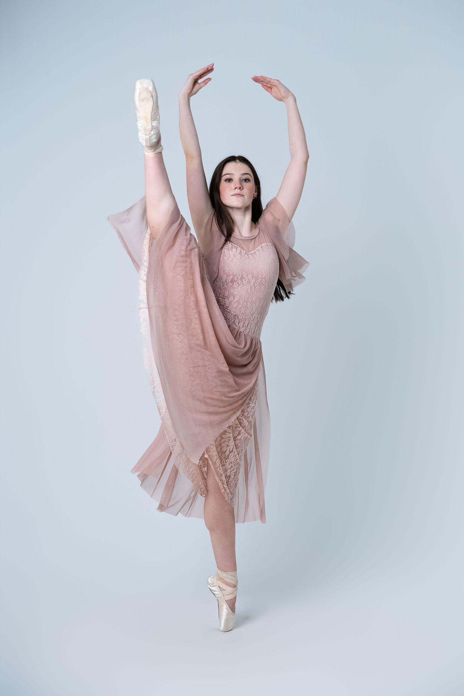 Ballet dancer in pink dress and pointe shoes performs elegant arabesque against grey backdrop.