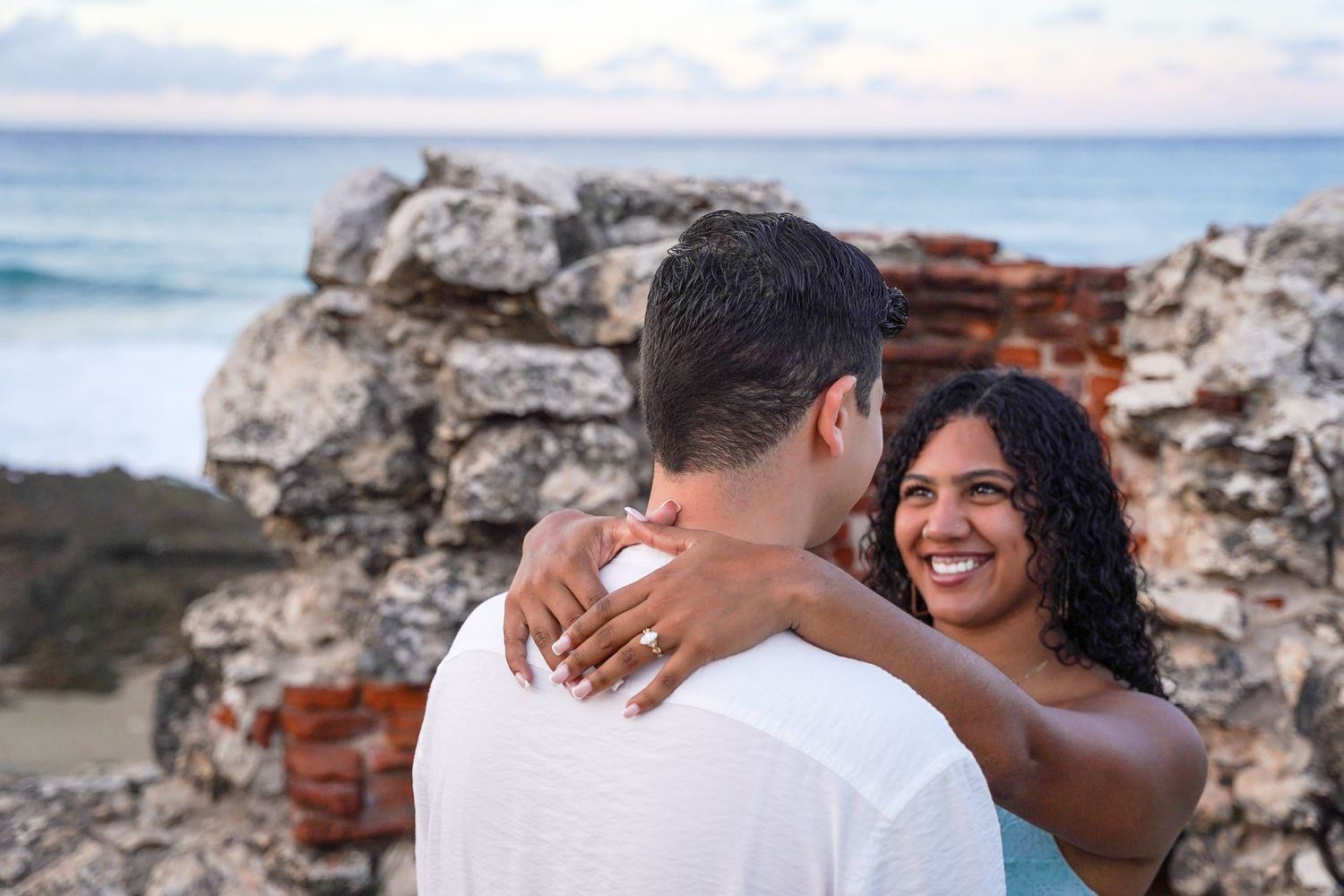 A newly engaged couple embracing on a beach with waves crashing against rocky cliffs in the background.