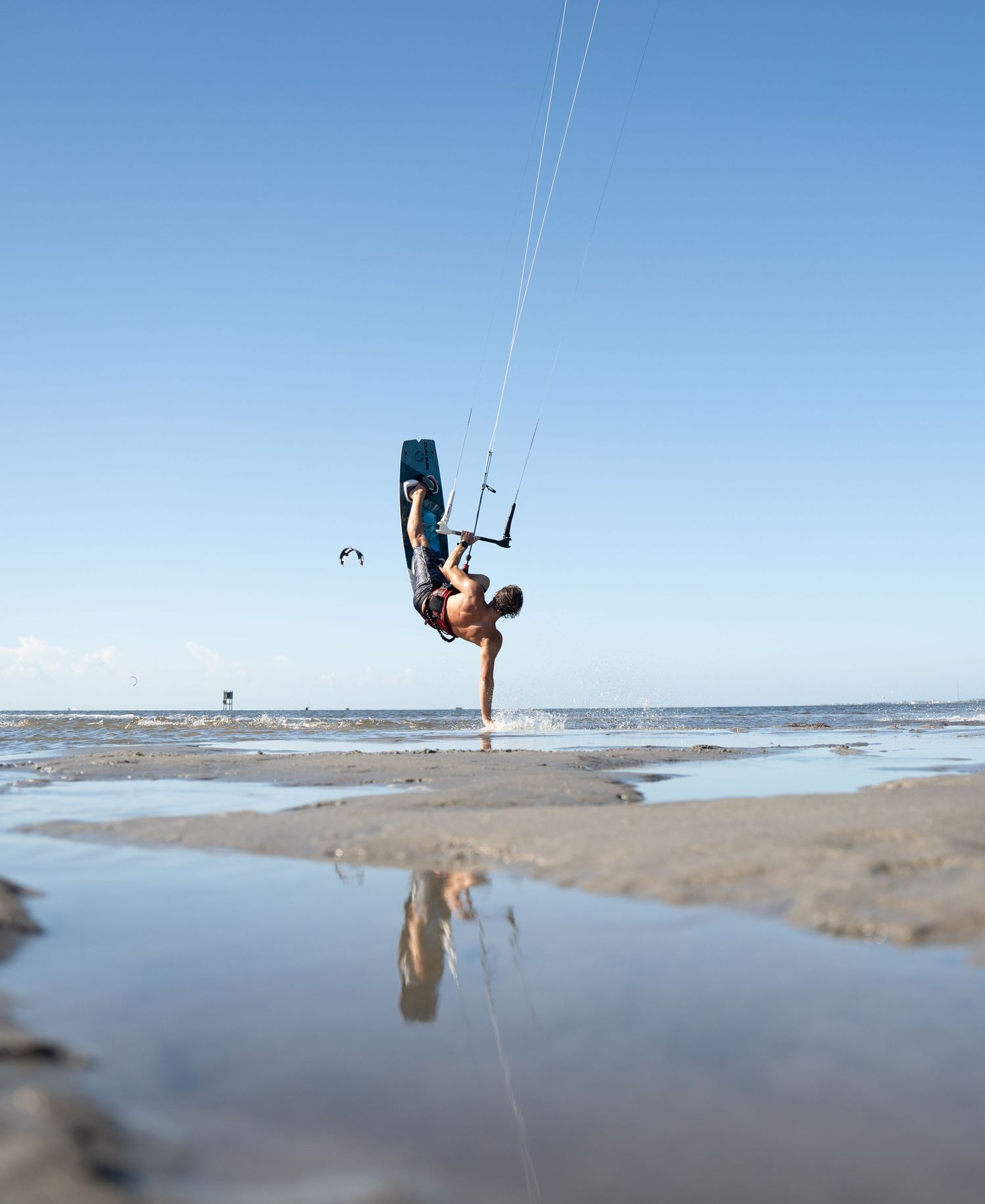 Kiteboarder performing an inverted trick on a beach with reflection in shallow water.