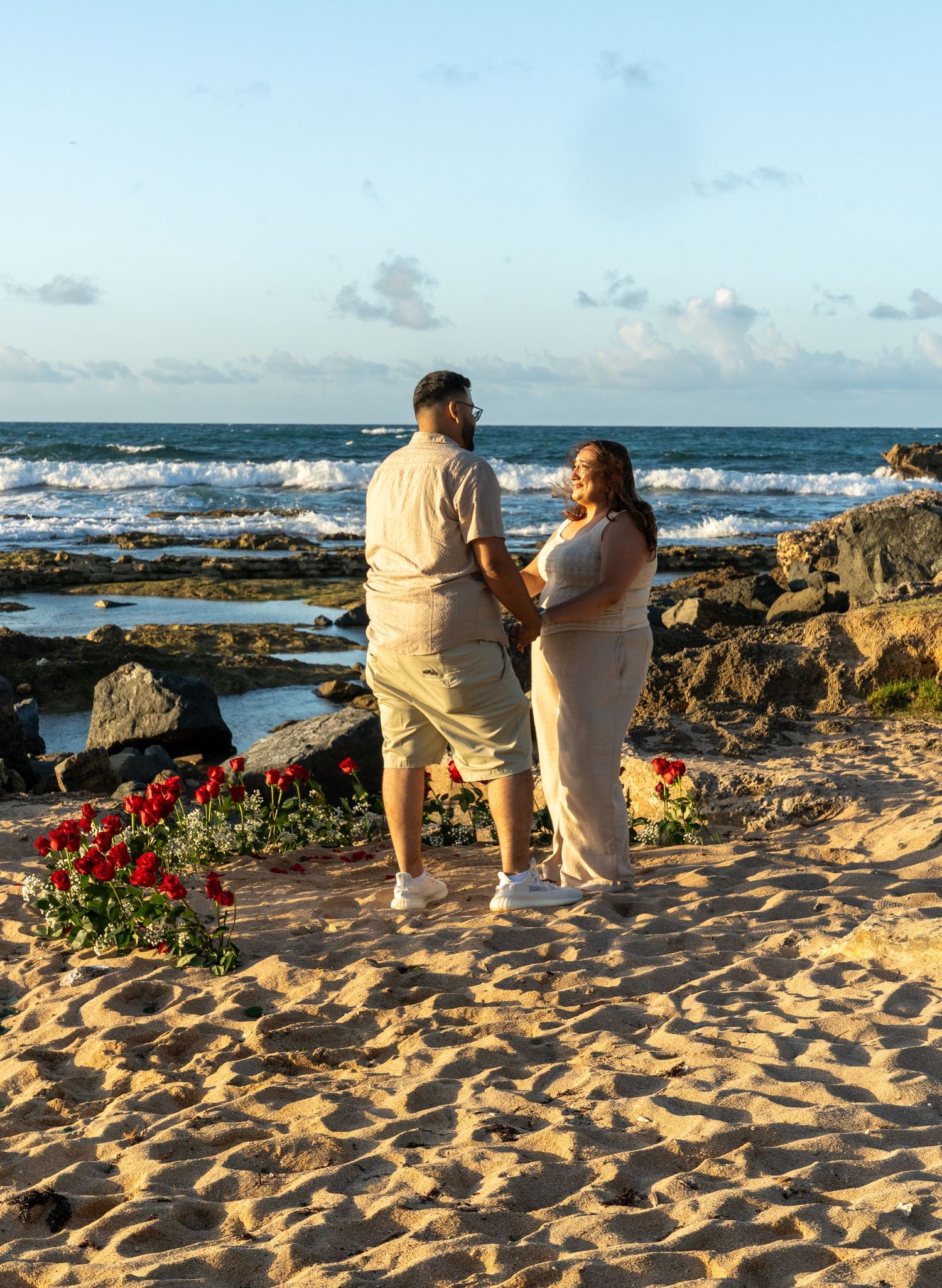 A couple stands together on a sandy beach with red flowers and ocean waves in the background.