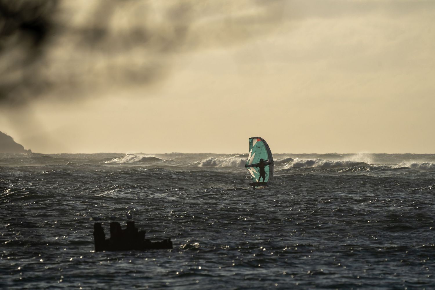 Distant wing foiler rides waves against a moody seascape at sunset.