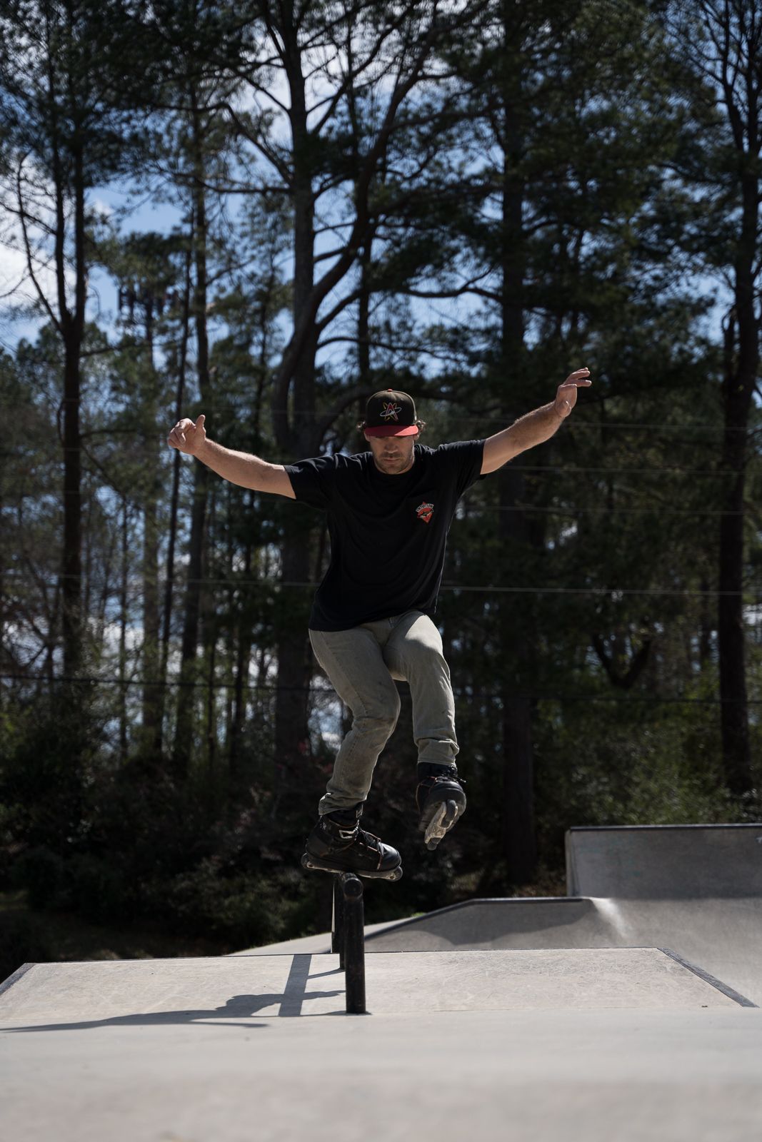 A skateboarder performs a trick on a rail at a skate park surrounded by pine trees.