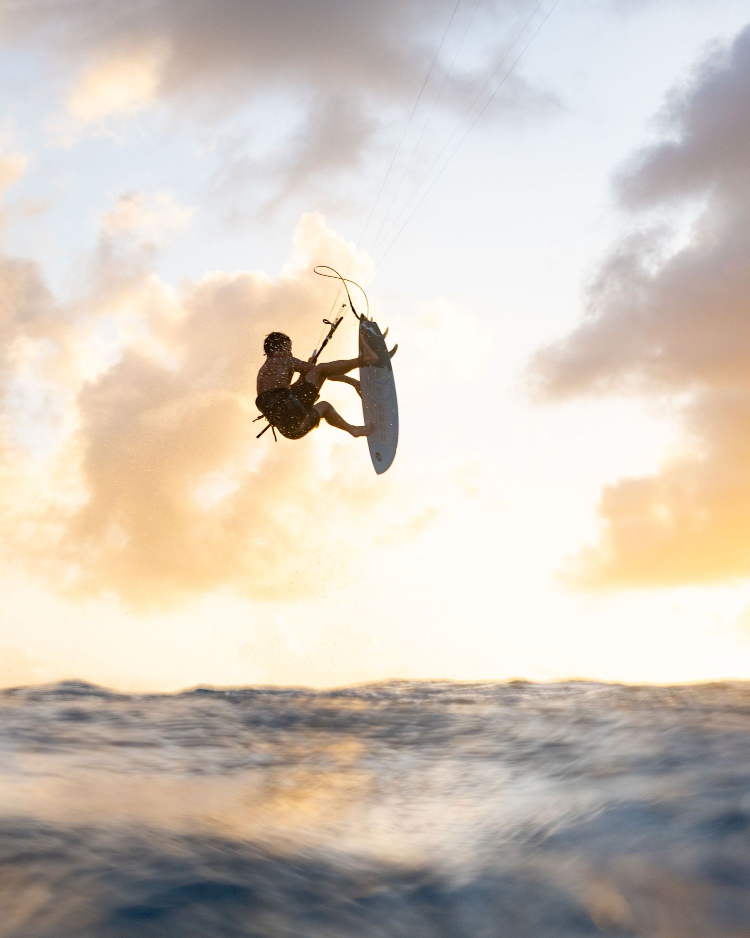 Silhouetted kitesurfer performs backflip trick against orange sunset sky.
