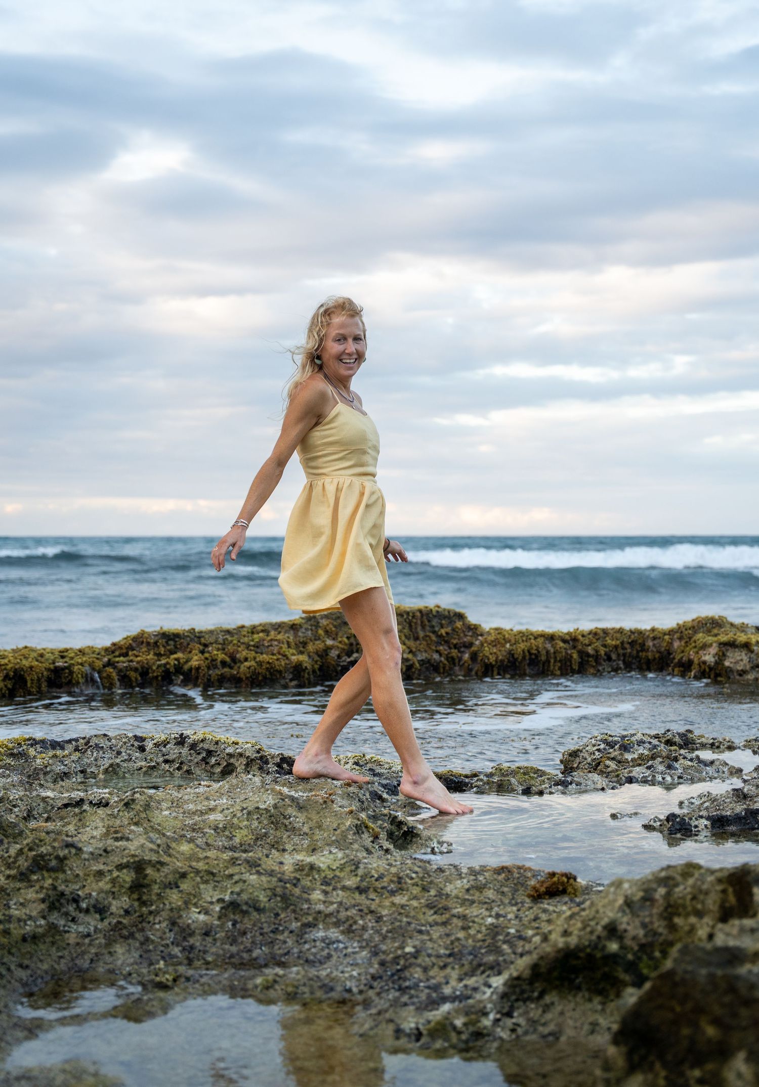 Person in yellow dress exploring rocky tide pools along a dramatic coastline during overcast weather. Portrait photography in San Juan