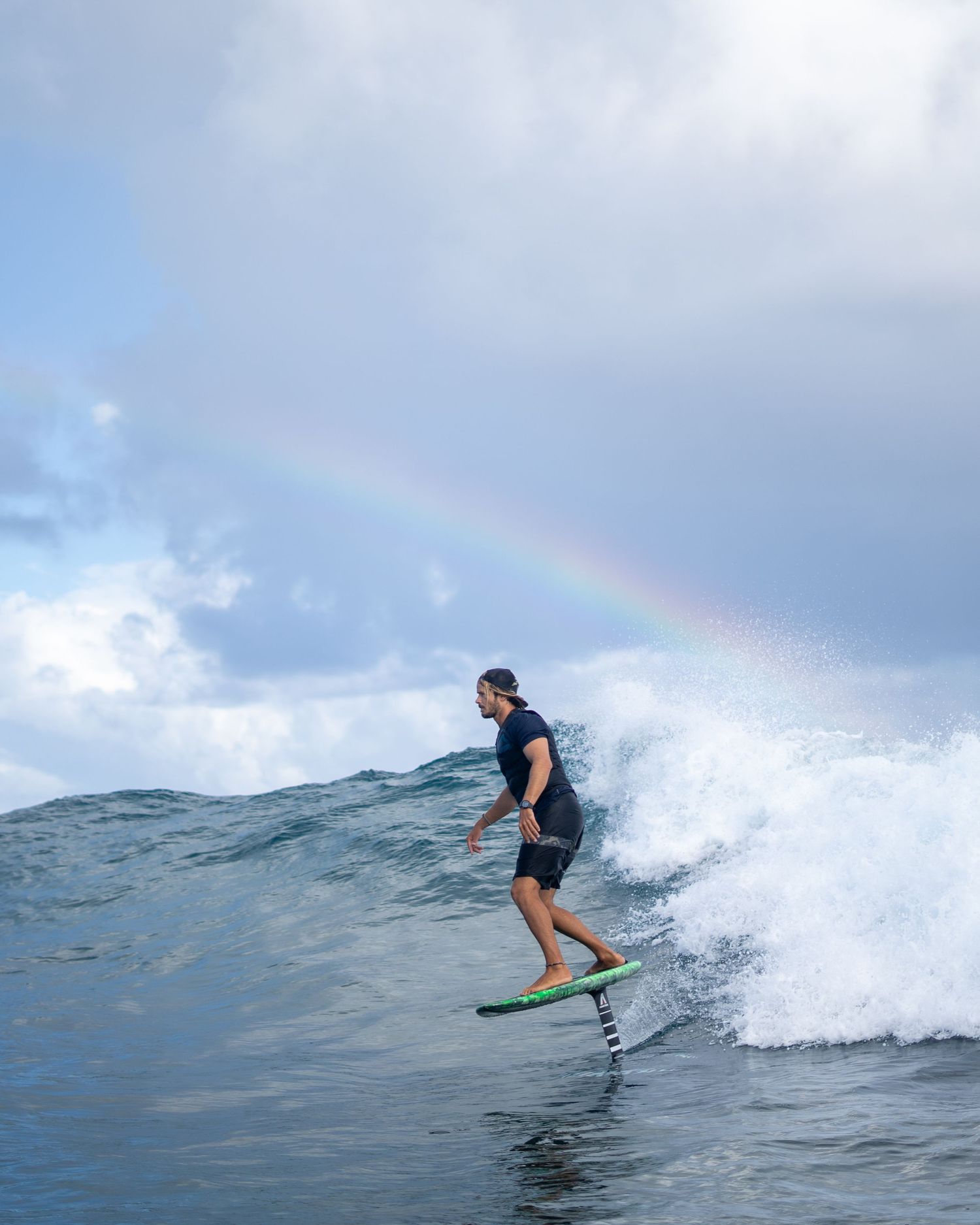 Foiler rides an ocean wave during sunrise with dramatic clouds and rainbow in background.
