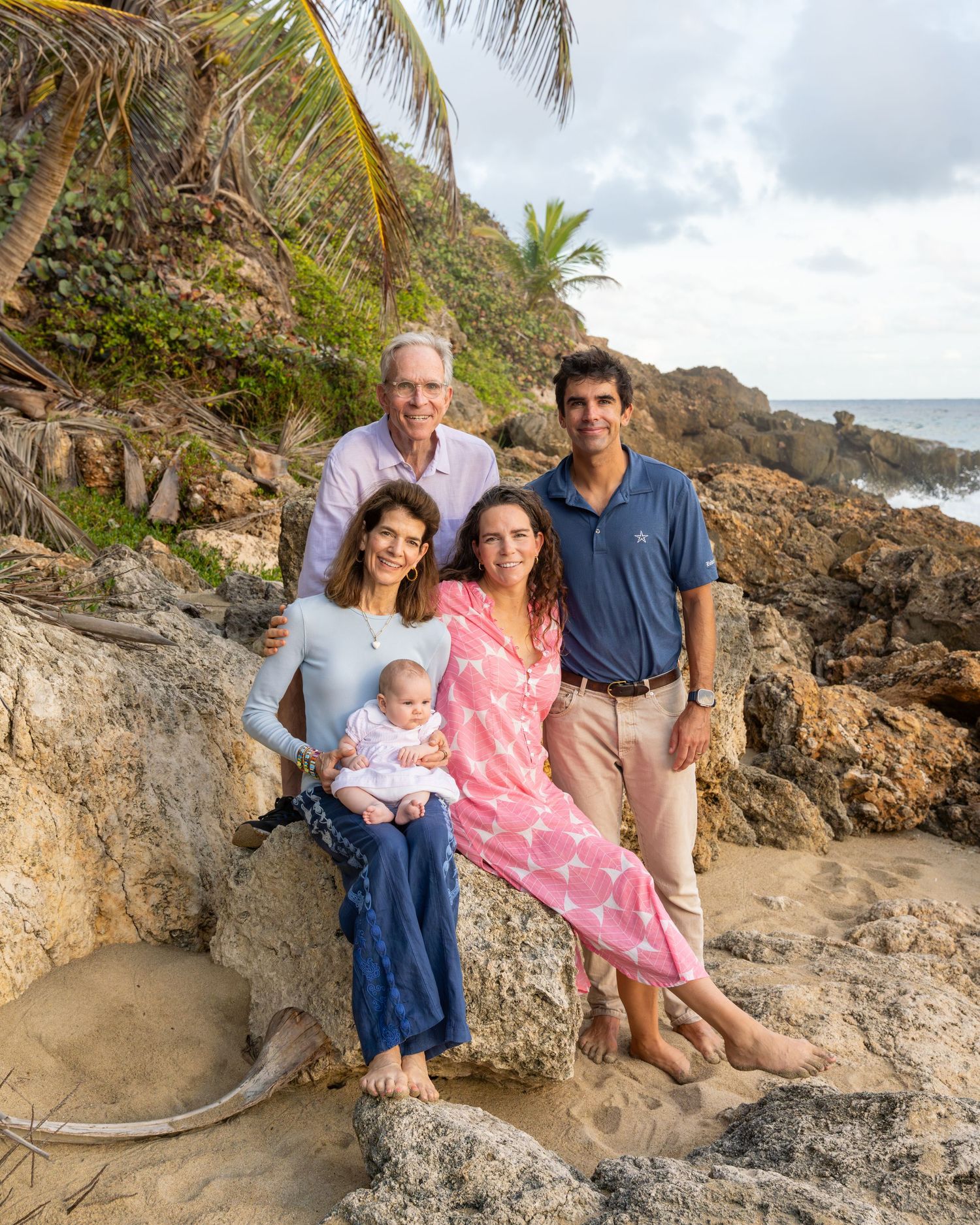 Family of five poses on rocky beach with lush palm trees and ocean waves in the background.