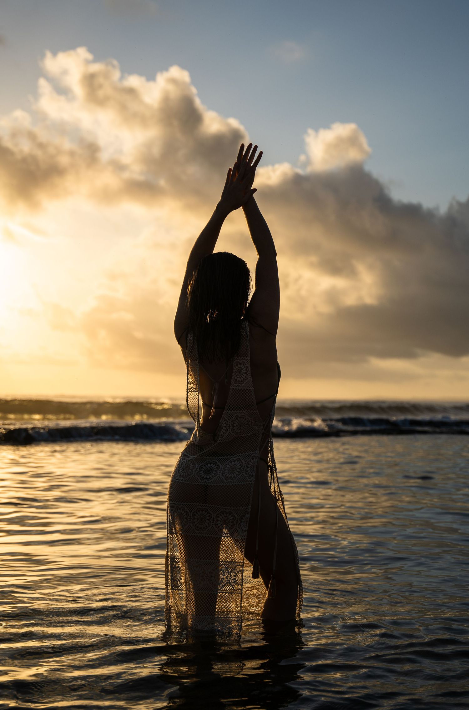 Silhouetted figure in water during golden sunset with dramatic clouds over ocean horizon.