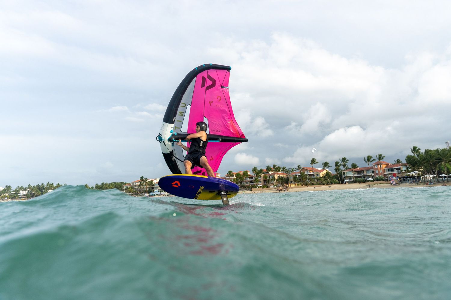 A pink windsurfing sail emerges from turquoise waters near a tropical coastline.