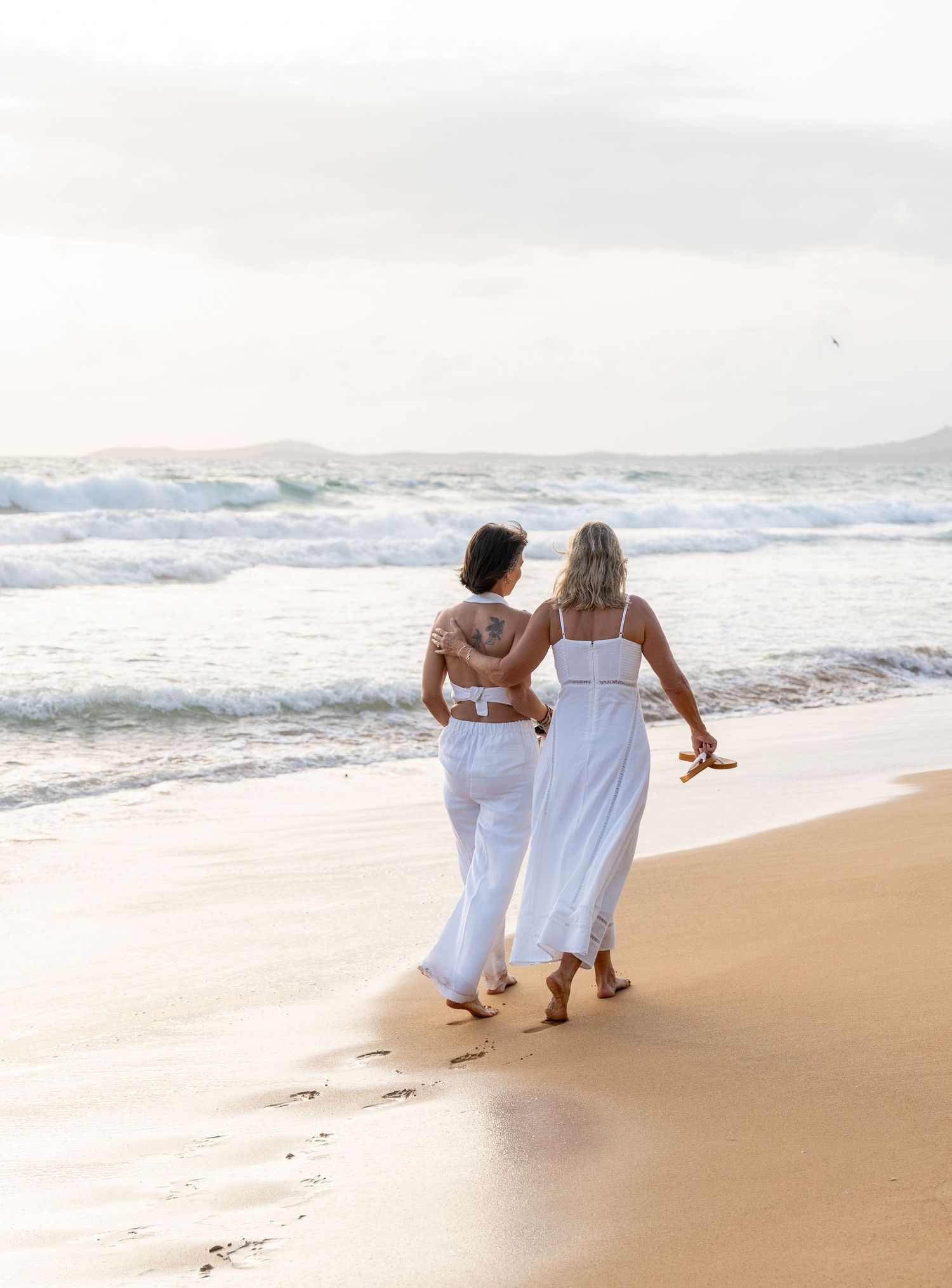 Two people in white clothing walk together along a sandy beach at sunset.