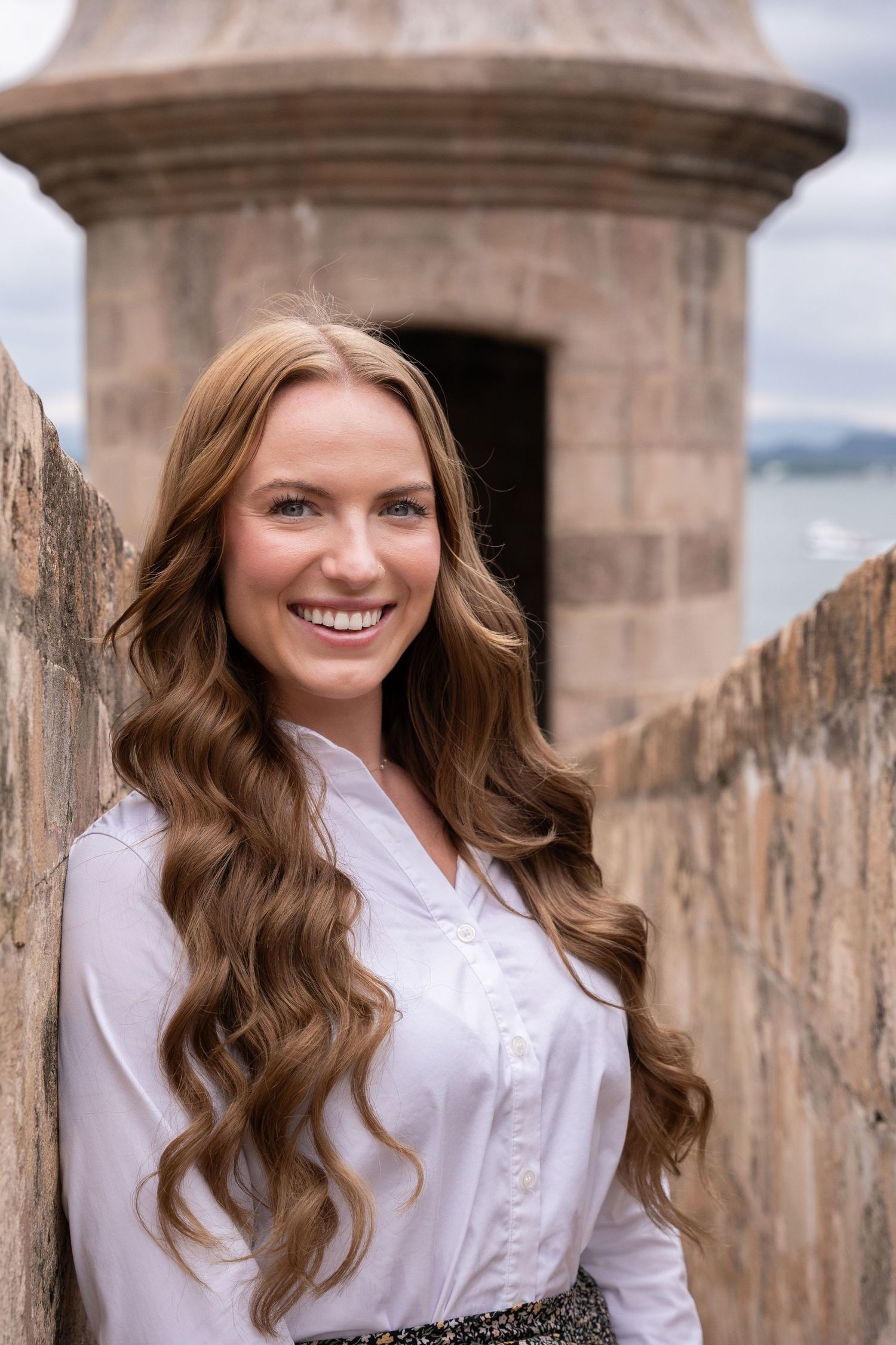 A portrait of someone with long wavy brown hair wearing a white button-up shirt against a stone wall backdrop.