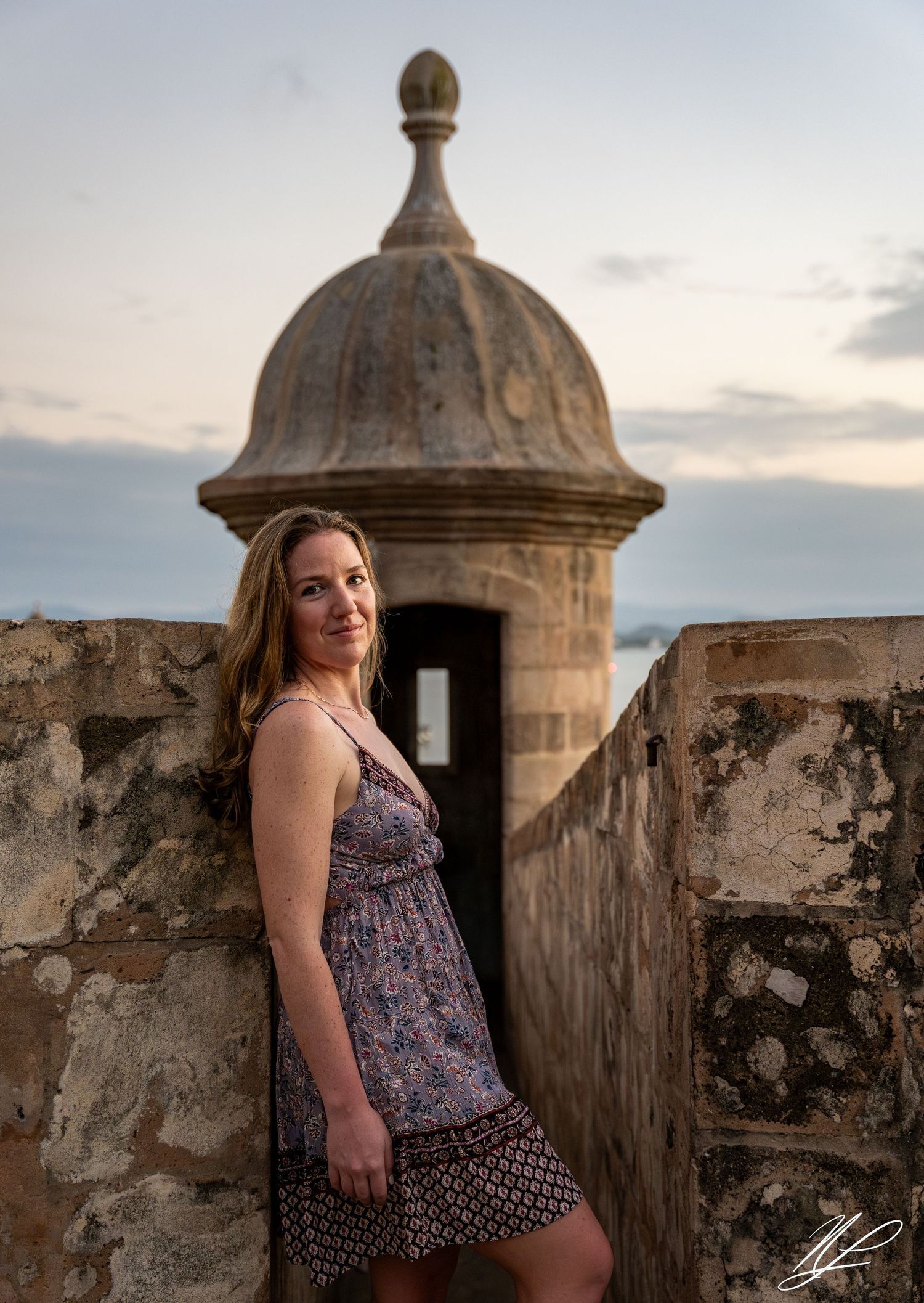 A woman in a floral dress poses against historic stone fortification walls at sunset. Portrait photography in Old San Juan