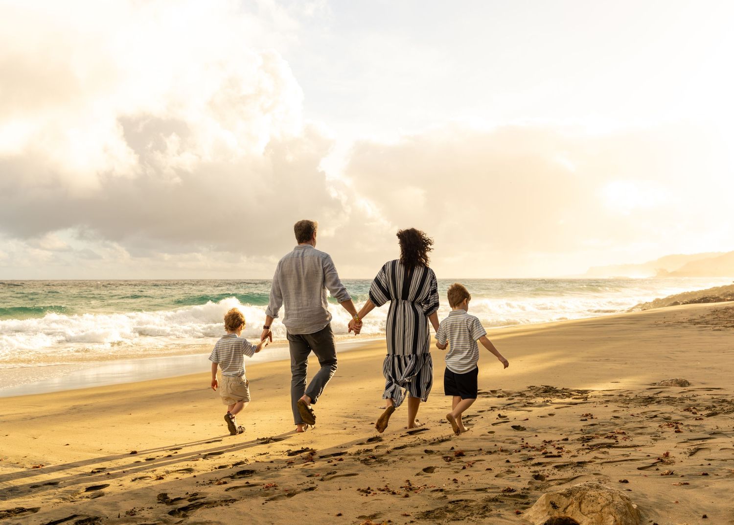 Family of four walking hand-in-hand along a sunny beach with ocean waves and golden sand.