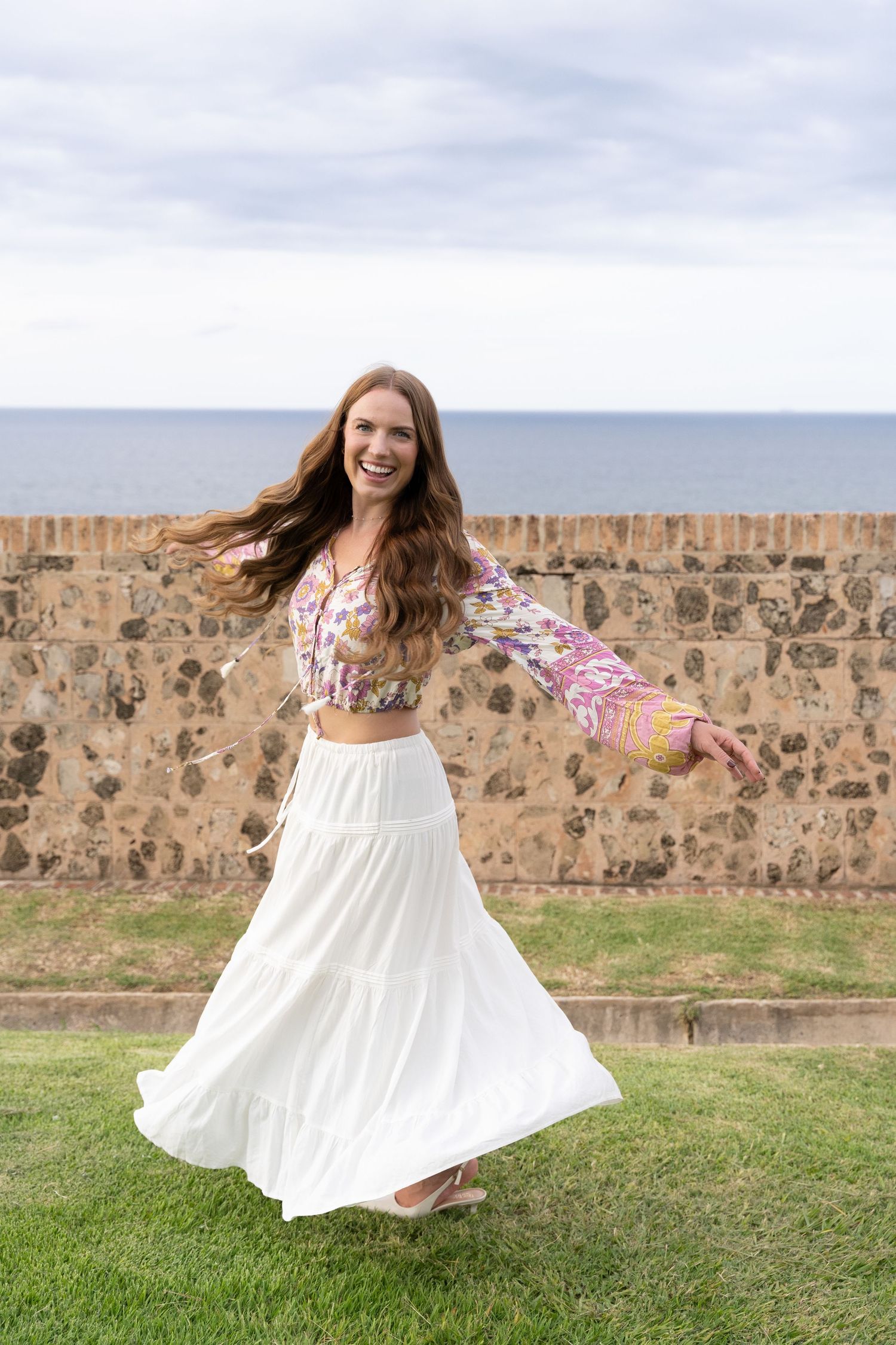 A person in a floral top and white skirt twirls joyfully near a stone wall by the ocean.